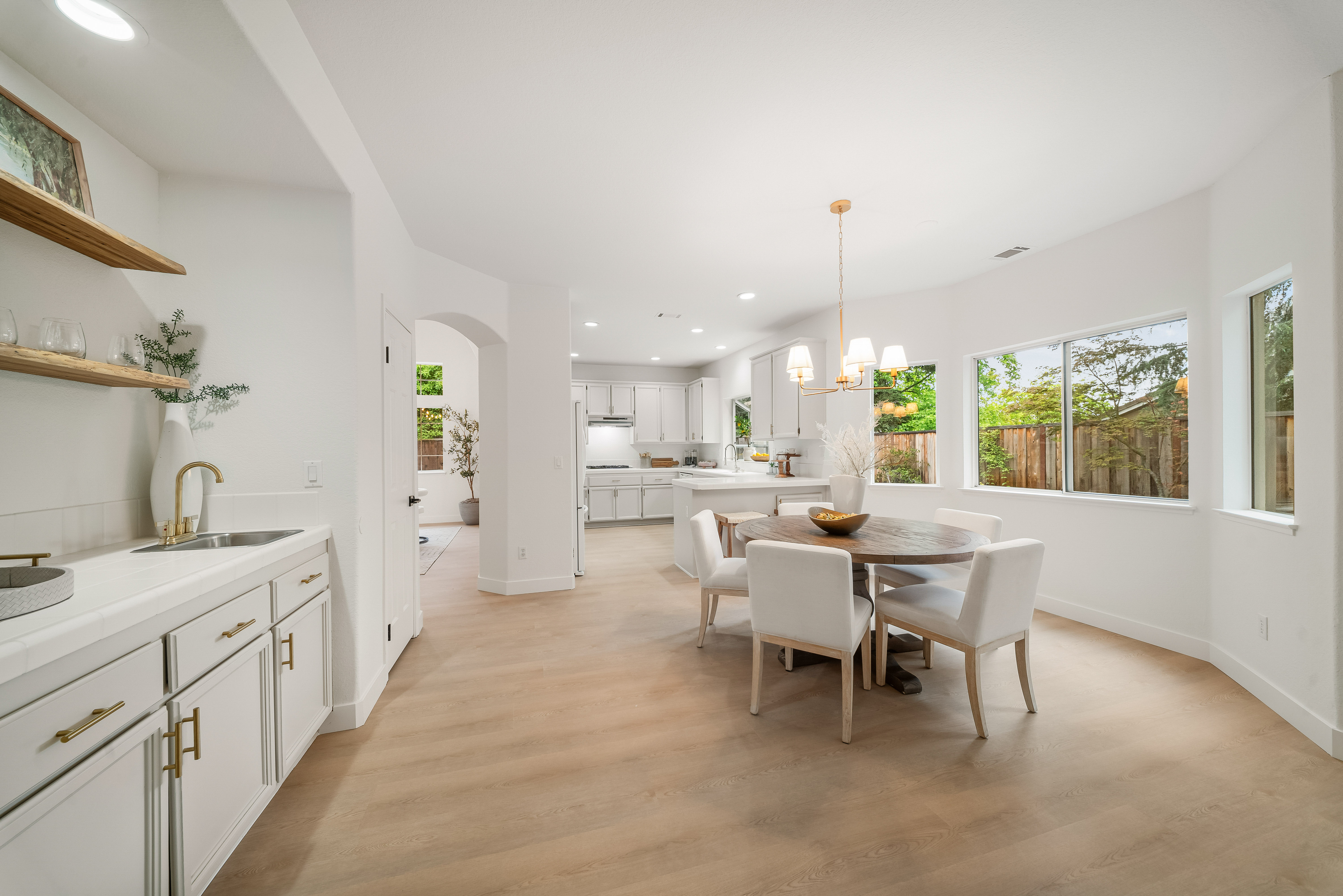 This bright, open-concept dining area features a round wooden table surrounded by four upholstered chairs, illuminated by a modern gold-toned chandelier. To the left, a built-in wet bar with white cabinetry, gold hardware, and open wood shelving provides a functional and stylish transition space. The room flows seamlessly into the adjacent kitchen, with light-toned wood flooring and large windows that invite ample natural light into the home.