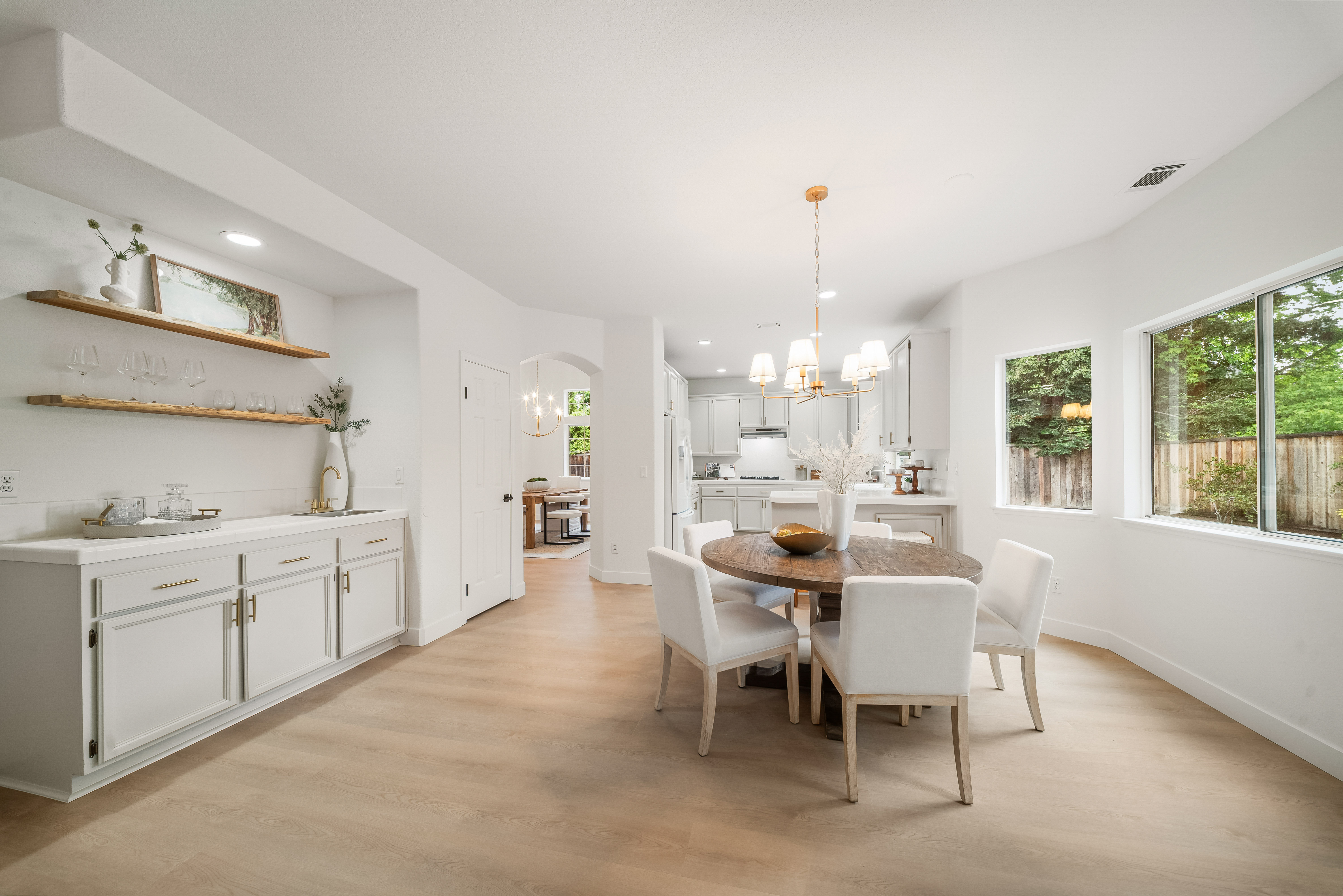 This bright and airy dining area features a round wooden table surrounded by four white upholstered chairs, illuminated by a modern gold-toned chandelier. To the left, a built-in wet bar with light-colored cabinetry and floating wood shelves provides a sophisticated touch, while light-toned wood flooring flows throughout the space. Large windows allow natural light to fill the room, creating an inviting atmosphere that transitions seamlessly into the adjacent kitchen.