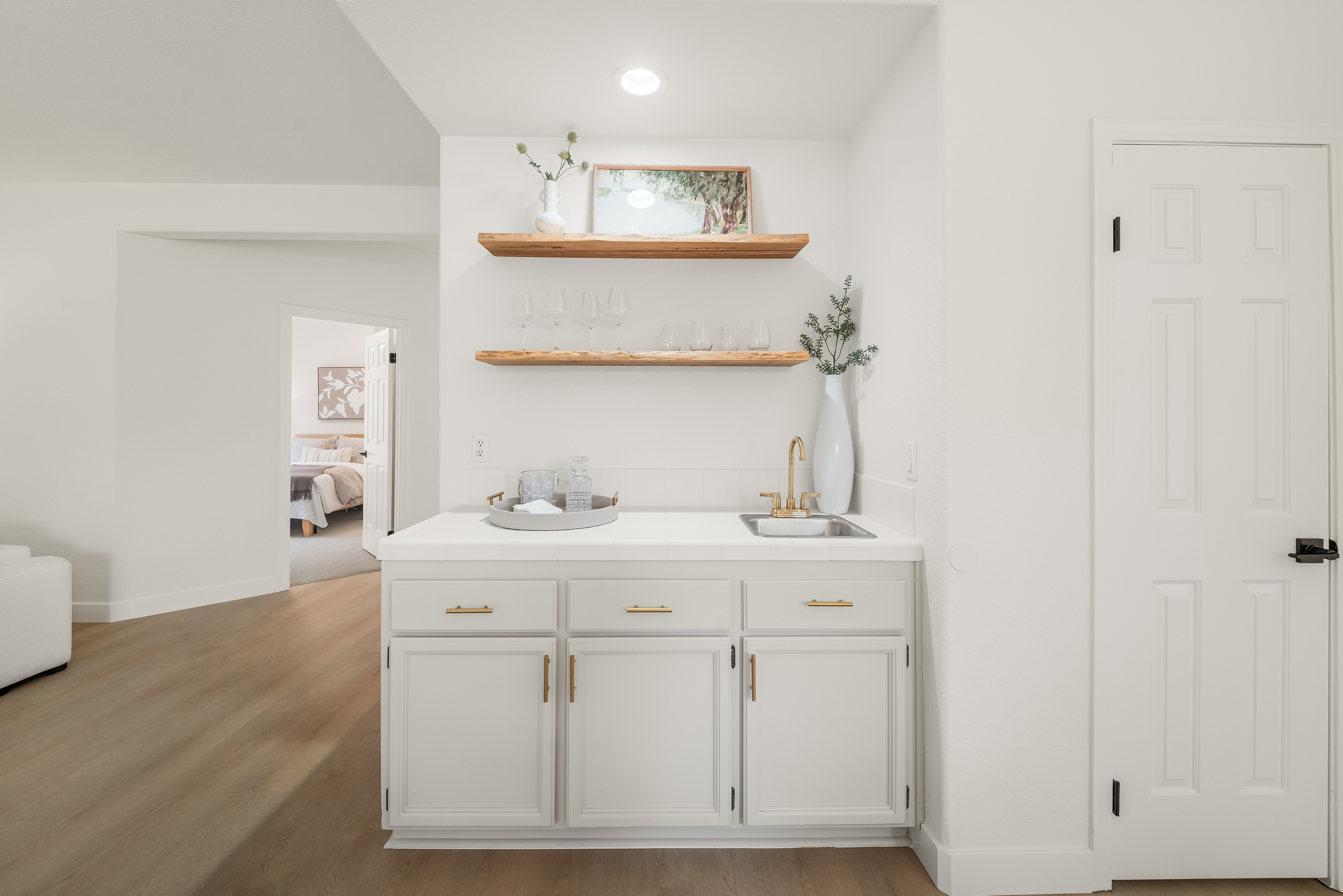 This interior view features a wet bar area with white cabinetry, gold-toned hardware, and a light-colored countertop with an integrated sink. Two natural wood floating shelves are mounted above the counter, displaying glassware and decorative items, while a tall white vase adds a touch of greenery. The space is bright and clean, with a view into an adjacent bedroom and a white door to the right, creating a functional and stylish transition area.