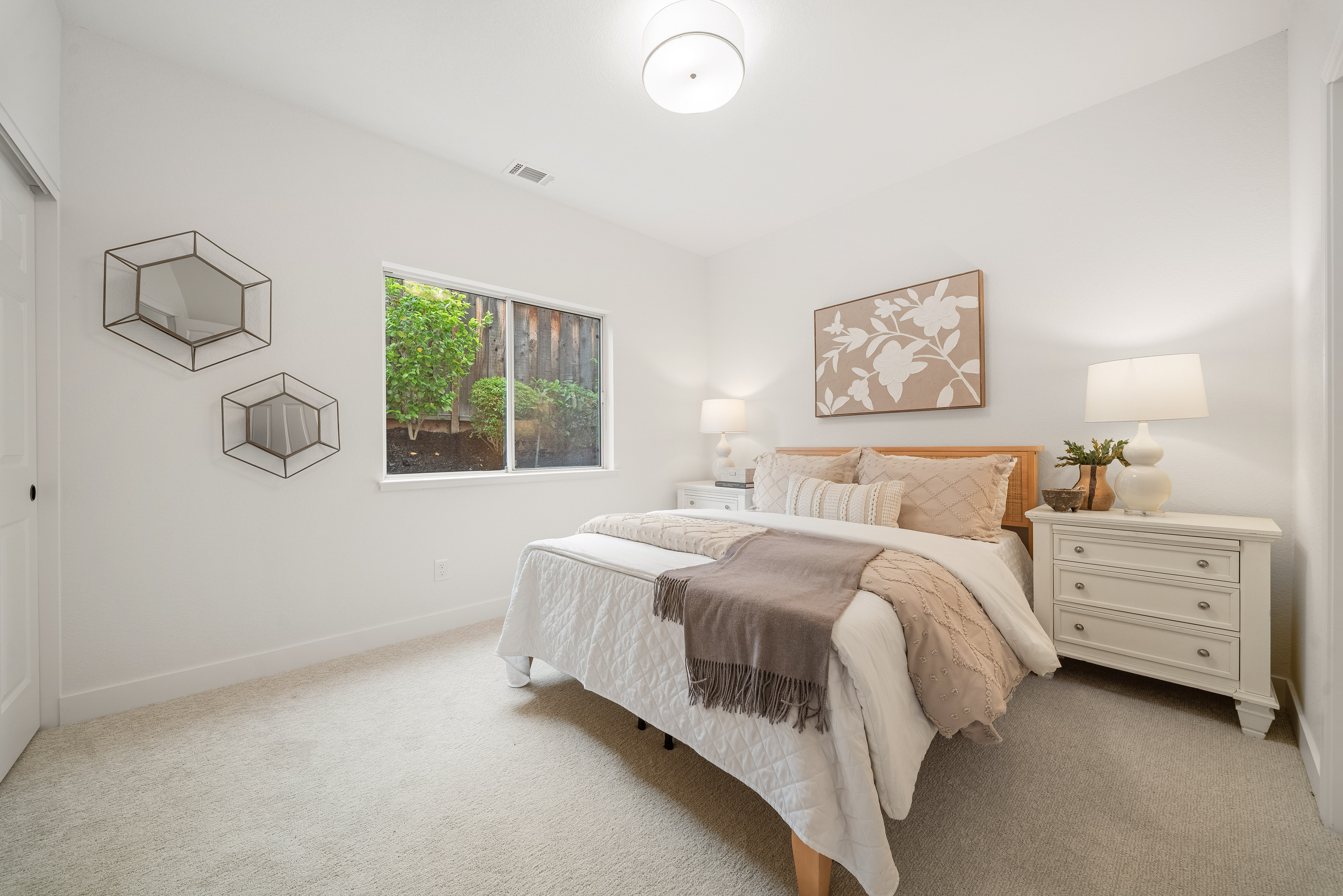 This bright and inviting bedroom features a neutral color palette with soft textures and light wood accents. The room is centered around a queen-sized bed dressed in layered bedding, flanked by a white nightstand and a matching dresser with a lamp. A window provides natural light and a view of the backyard, while geometric wall mirrors add a modern decorative touch.
