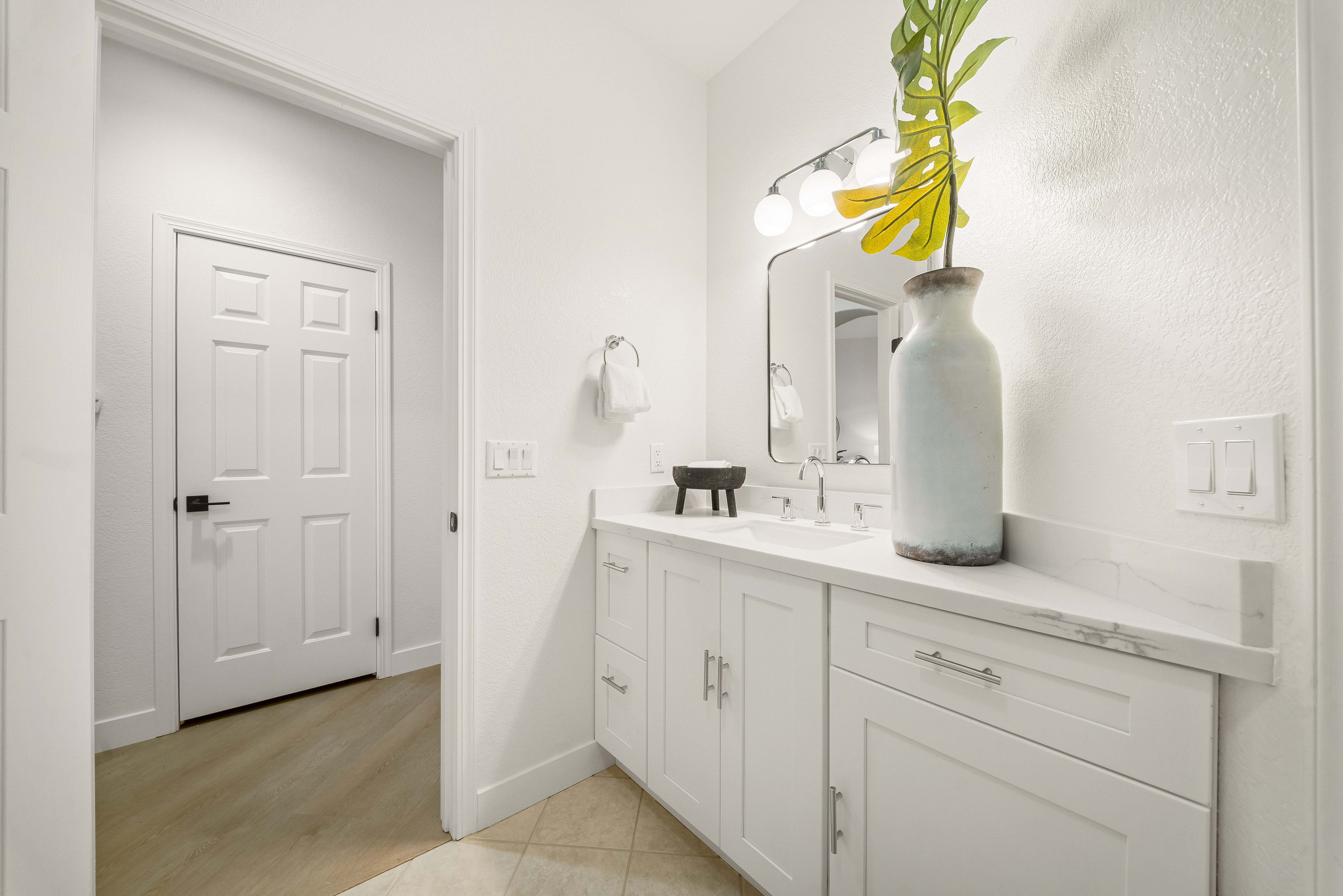 This bright and clean bathroom features a white vanity with a stone countertop and a rectangular undermount sink. A large mirror is mounted above the vanity, complemented by a three-light fixture and a decorative vase with greenery. The space includes a white door leading to an adjacent room, with neutral-toned tile flooring and crisp white walls creating a modern, airy aesthetic.
