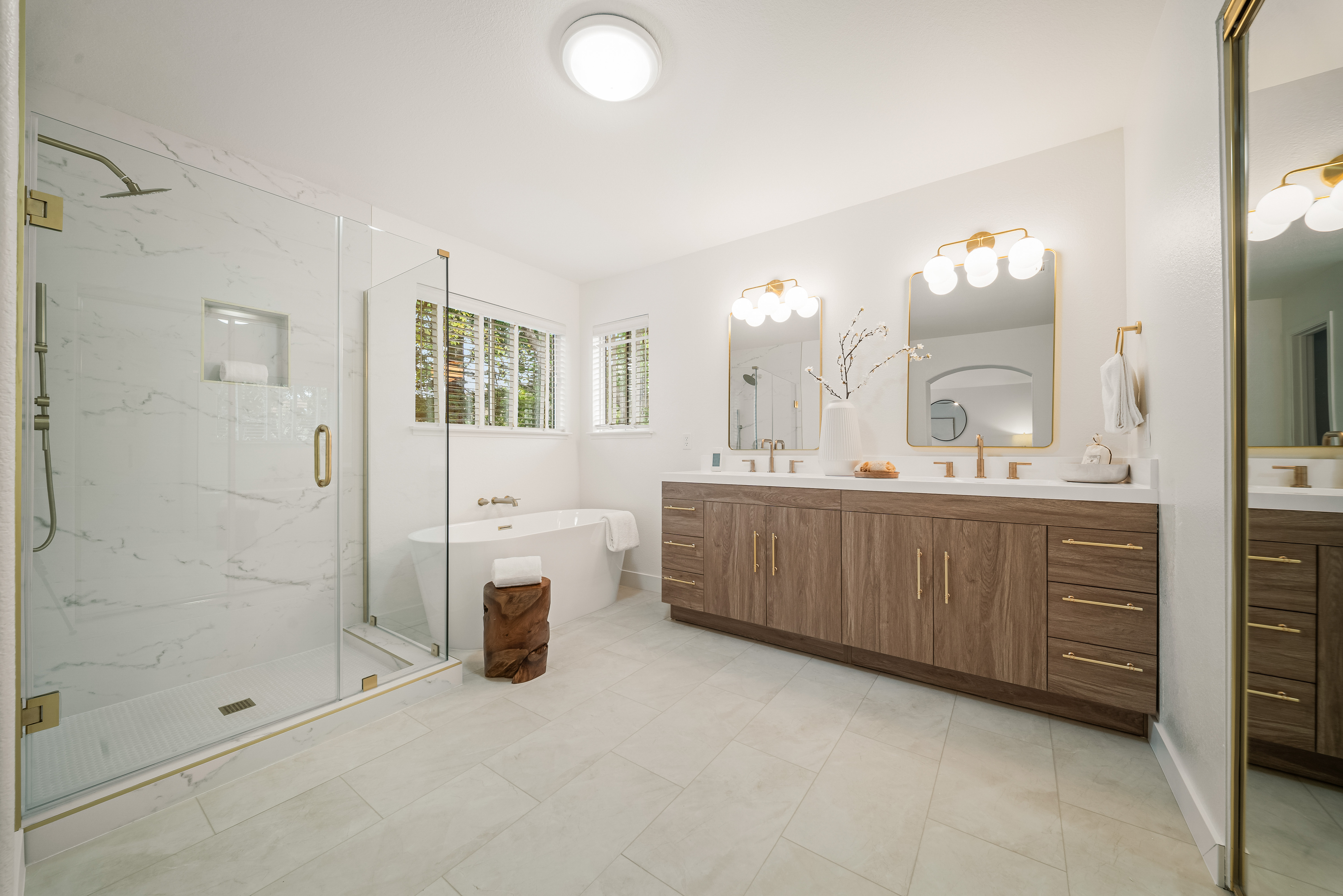 This bright and spacious primary bathroom features a double vanity with wood-toned cabinetry, gold-toned hardware, and dual mirrors illuminated by globe-style wall sconces. To the left, a glass-enclosed walk-in shower with light-colored stone tile walls sits beside a freestanding soaking tub positioned under a window. The room is finished with light-colored floor tiles, creating a clean, modern, and luxurious atmosphere.