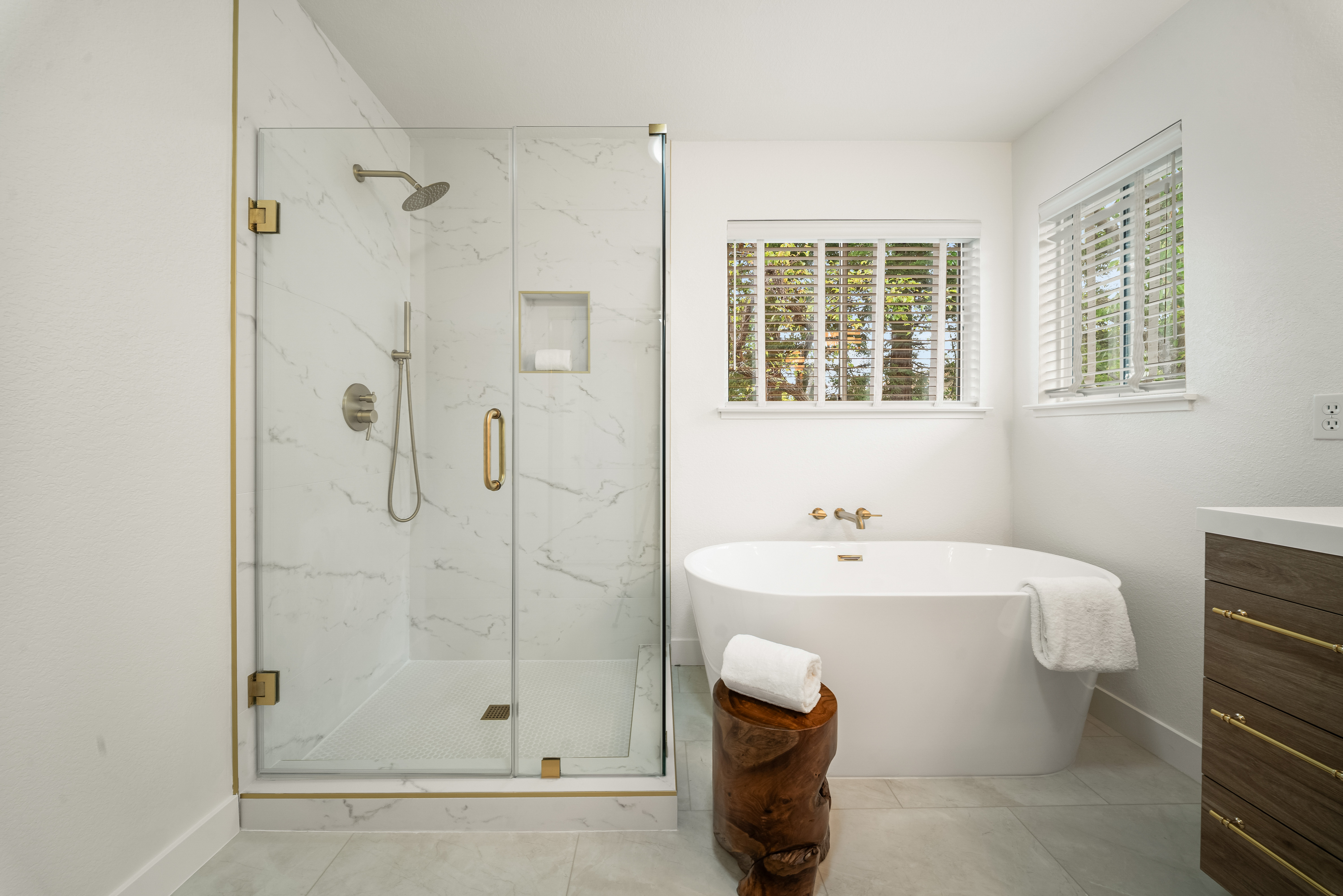 This bright primary bathroom features a glass-enclosed walk-in shower with patterned wall tile and a freestanding soaking tub positioned beneath a window. A rustic wooden stool sits in the foreground, adding a natural texture to the clean, modern aesthetic. The space is finished with gold-toned fixtures and a dark wood vanity, creating a sophisticated and spa-like atmosphere.