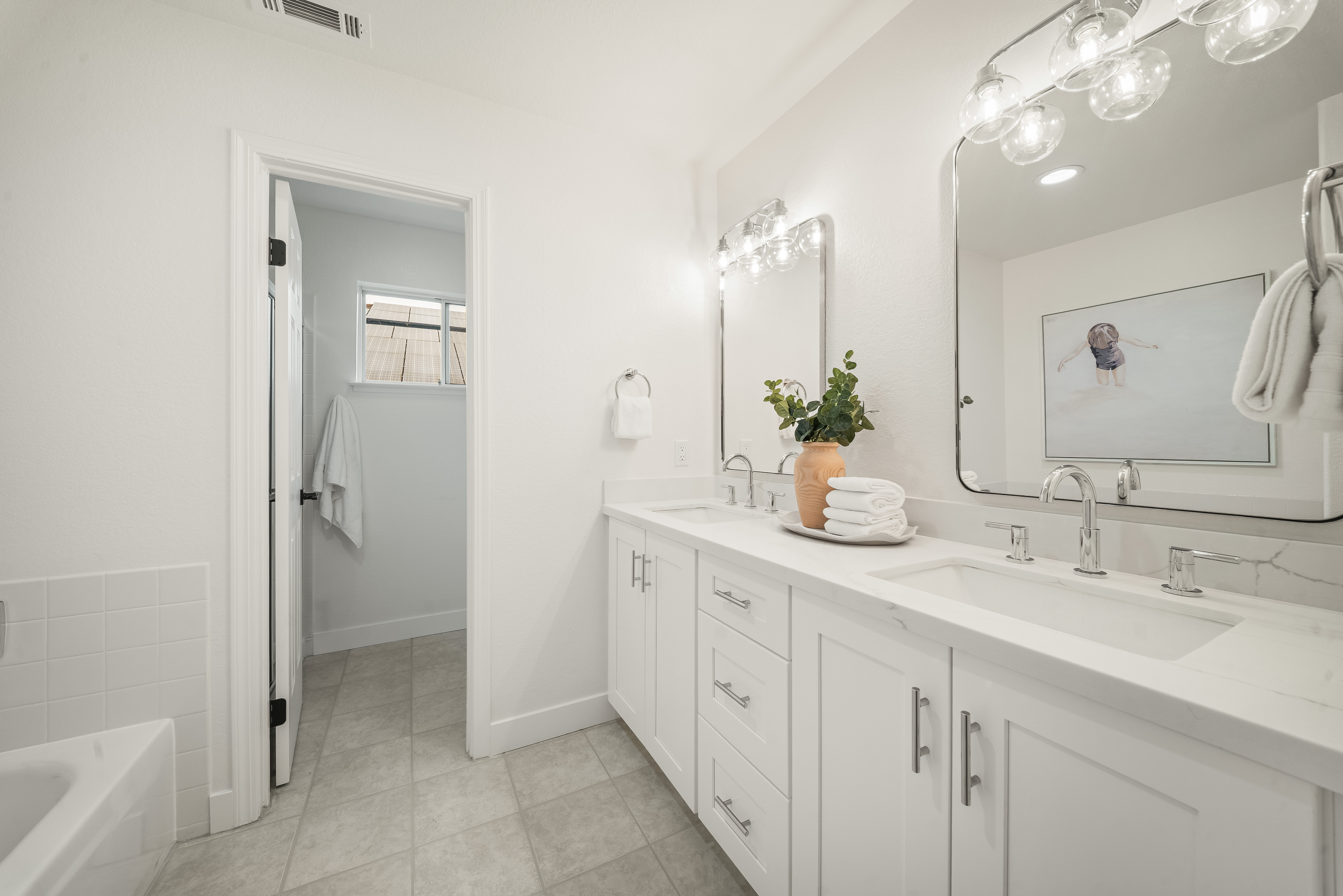 This bright and clean bathroom features a double vanity with white cabinetry, light-toned stone countertops, and dual rectangular sinks. The space is accented by polished chrome fixtures, two large mirrors with rounded corners, and a decorative vase with greenery. A doorway leads to an adjacent room with a window, while the neutral color palette and soft lighting create a serene and inviting atmosphere.