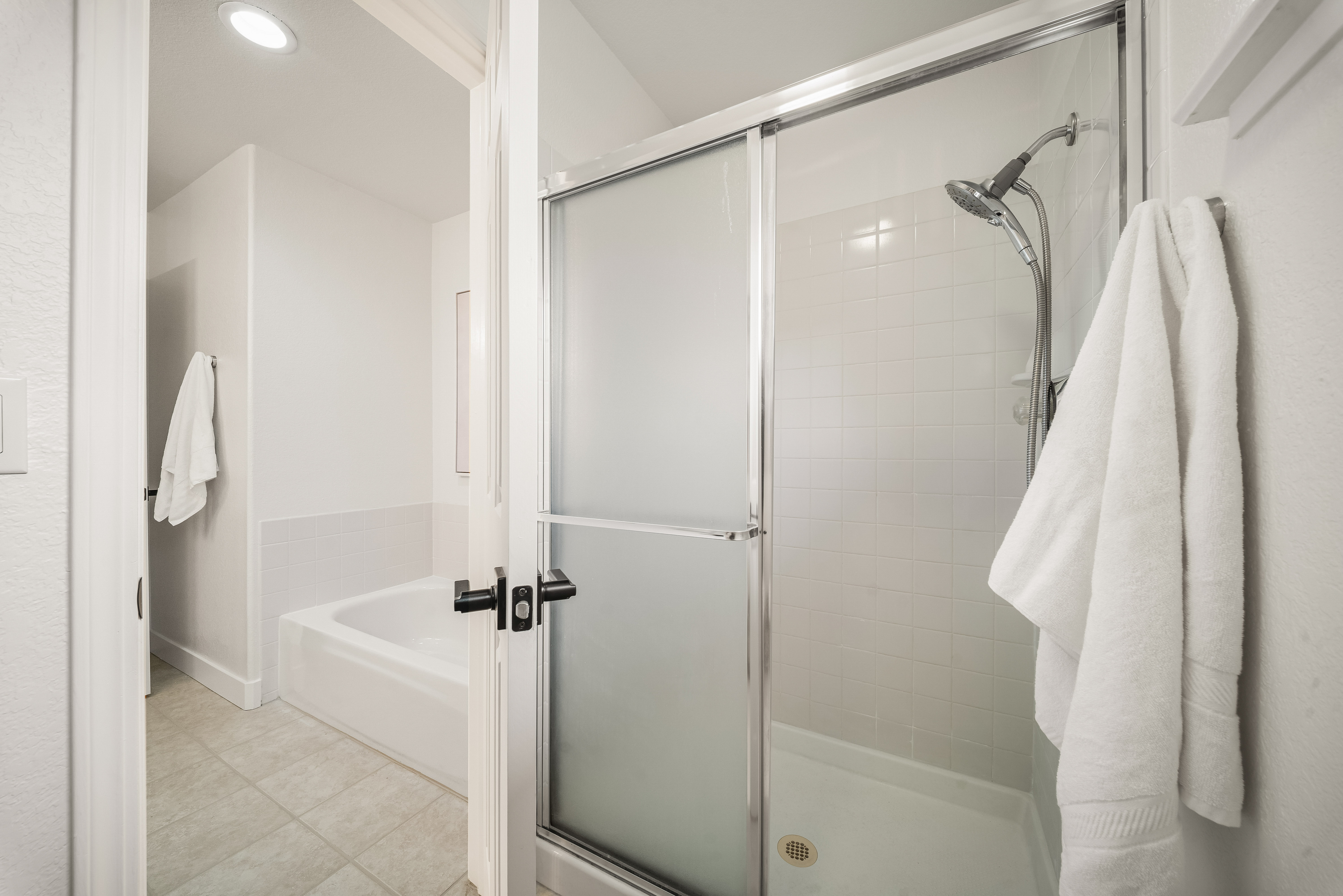 This bathroom features a walk-in shower with frosted glass doors and a handheld showerhead, adjacent to a separate soaking tub visible through the doorway. The space is finished with neutral-toned wall tiles and light-colored flooring, creating a clean and functional aesthetic. A white towel hangs on a rack near the shower, adding a touch of convenience to the bright, well-lit layout.