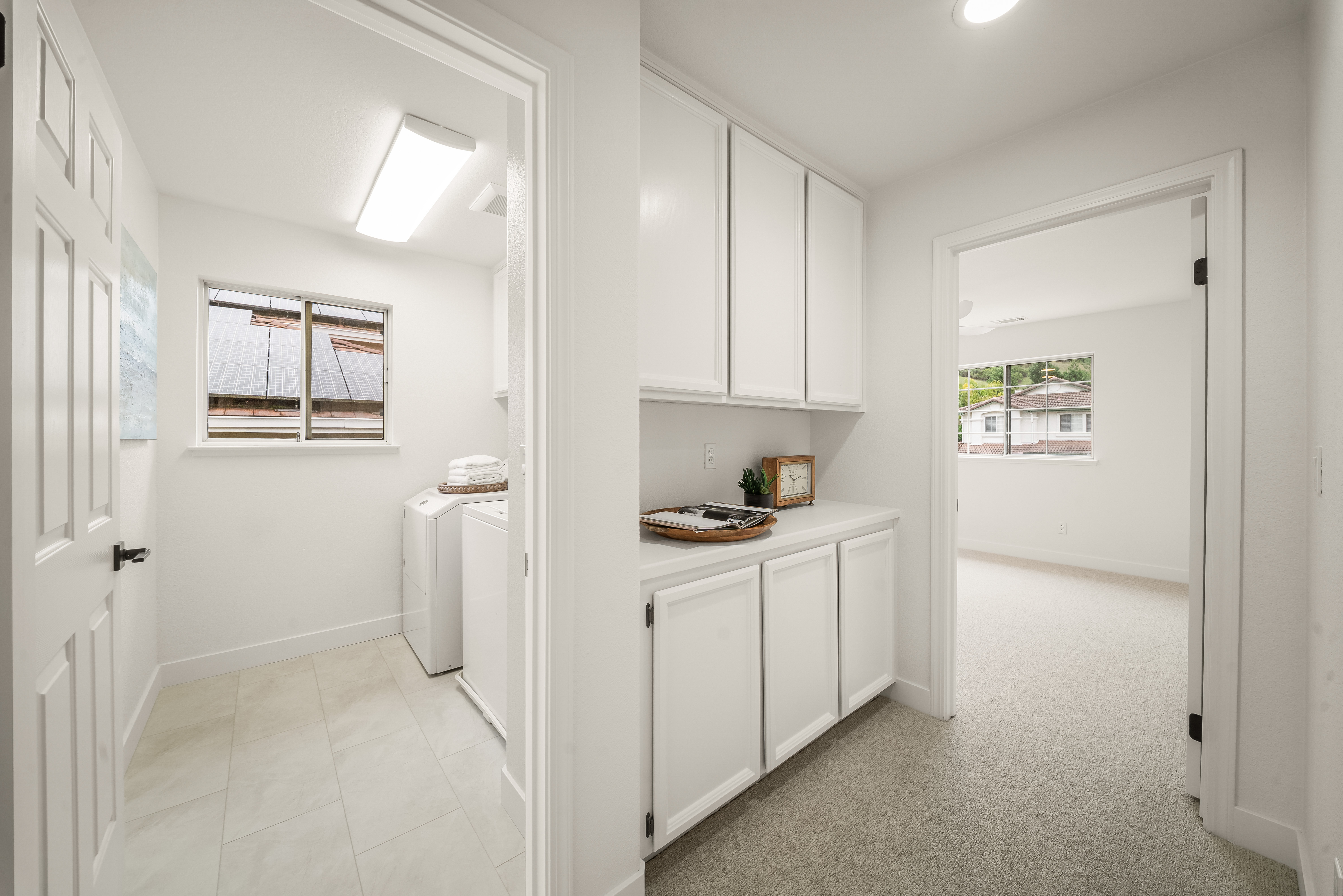 This view captures a functional laundry room featuring white appliances and tile flooring, connected to a hallway with built-in white cabinetry and a countertop. The perspective looks through a doorway into the laundry space on the left and into a bright, carpeted bedroom on the right. The neutral color palette and clean lines create a bright, organized, and cohesive transition between these living areas.