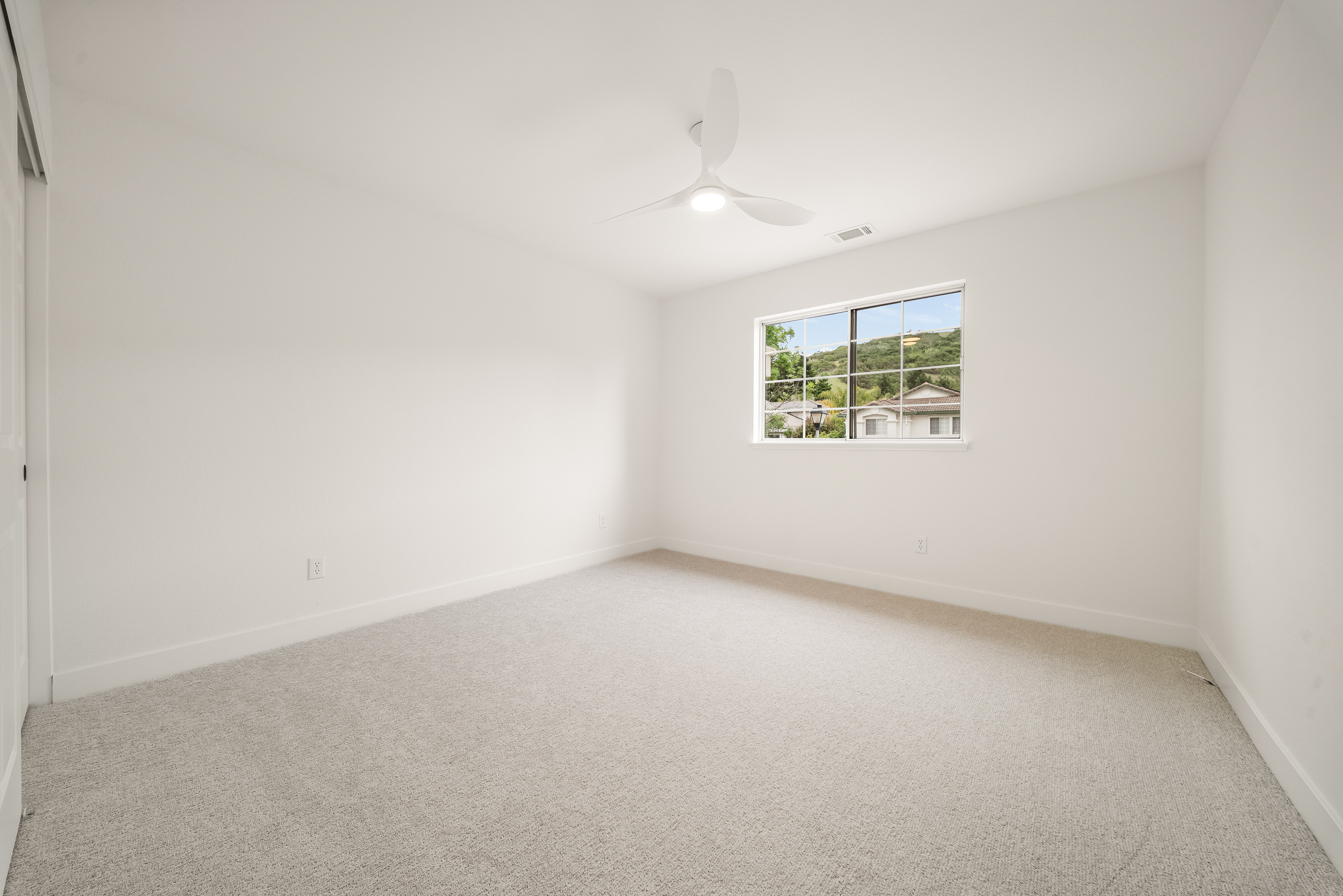 This is a bright, minimalist bedroom featuring neutral-toned carpeting and crisp white walls. A modern ceiling fan with integrated lighting is centered in the room, while a single window provides natural light and a view of the neighborhood. The space is clean and unfurnished, offering a versatile blank canvas for potential residents.