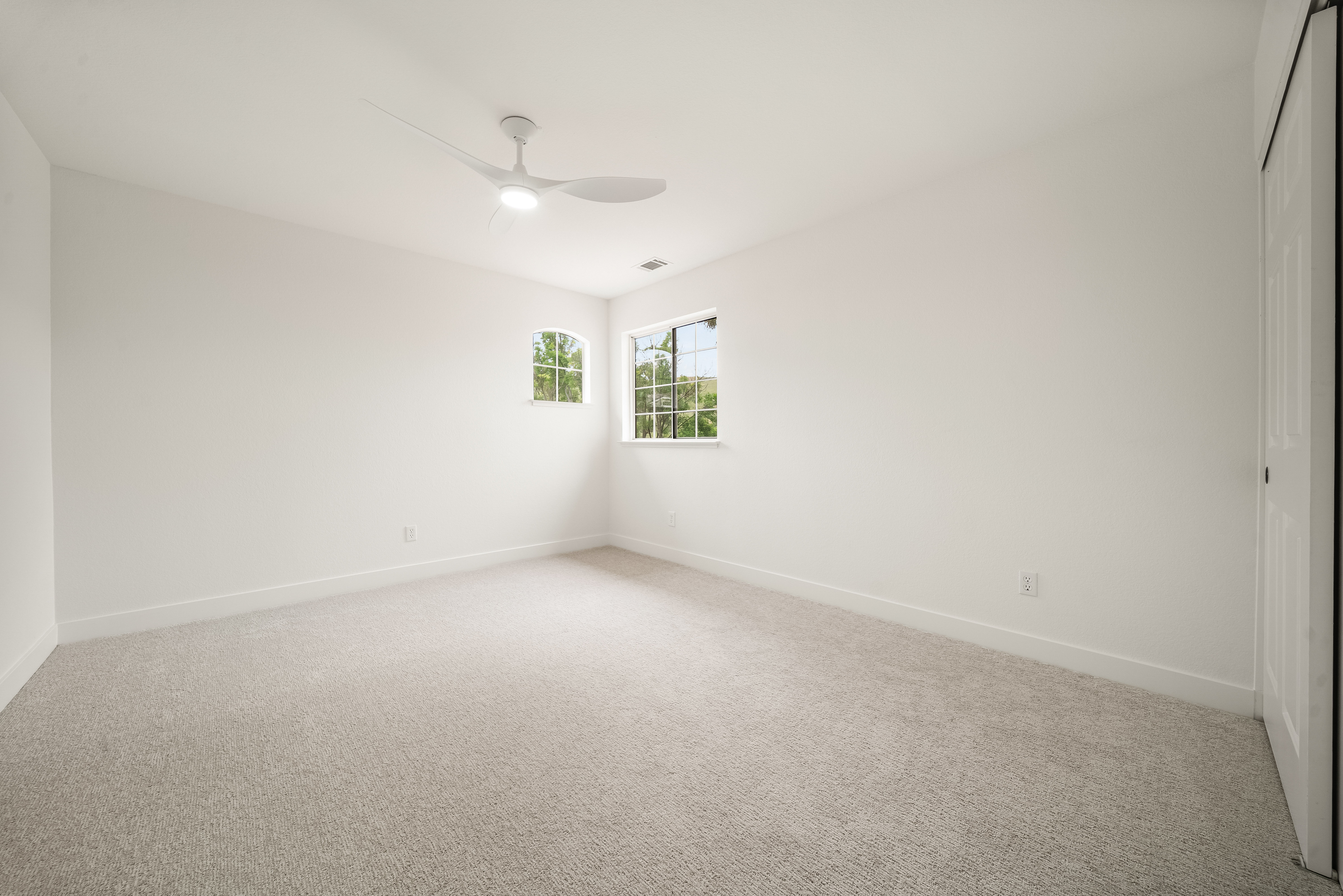 This bright, minimalist bedroom features neutral-toned carpeting and crisp white walls, creating a clean and versatile living space. The room is illuminated by a modern ceiling fan with an integrated light fixture and natural light streaming through two windows, including a unique arched window. A white closet door is visible on the right, completing the simple and functional layout of this room.
