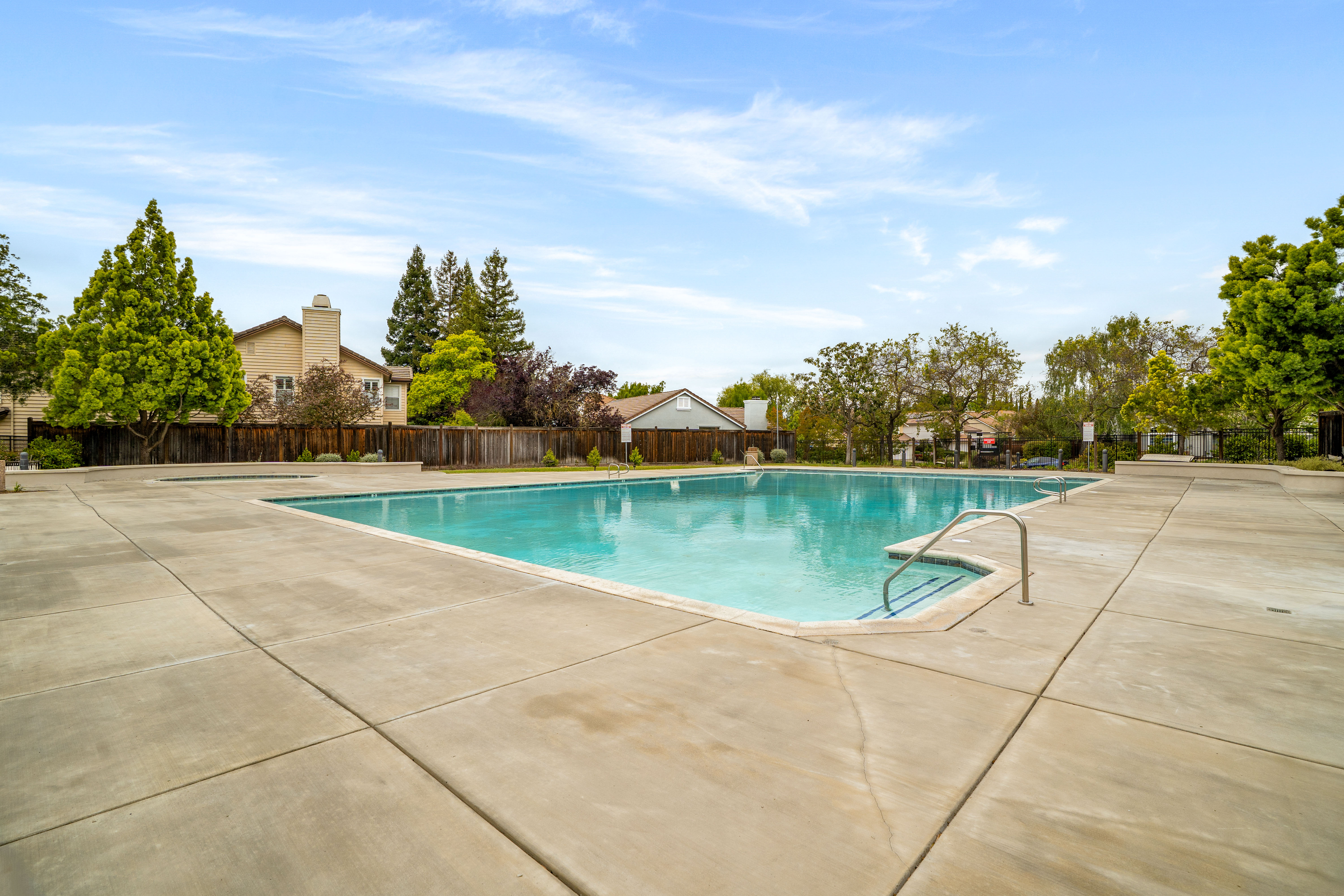 This image captures a spacious, rectangular community swimming pool set within a large, light-colored concrete deck. The pool features a set of metal handrails leading into the shallow end, with a clear blue water surface reflecting the bright, partly cloudy sky. The area is enclosed by a wooden fence and surrounded by mature trees and neighboring residential homes, creating a quiet and well-maintained outdoor recreational space.