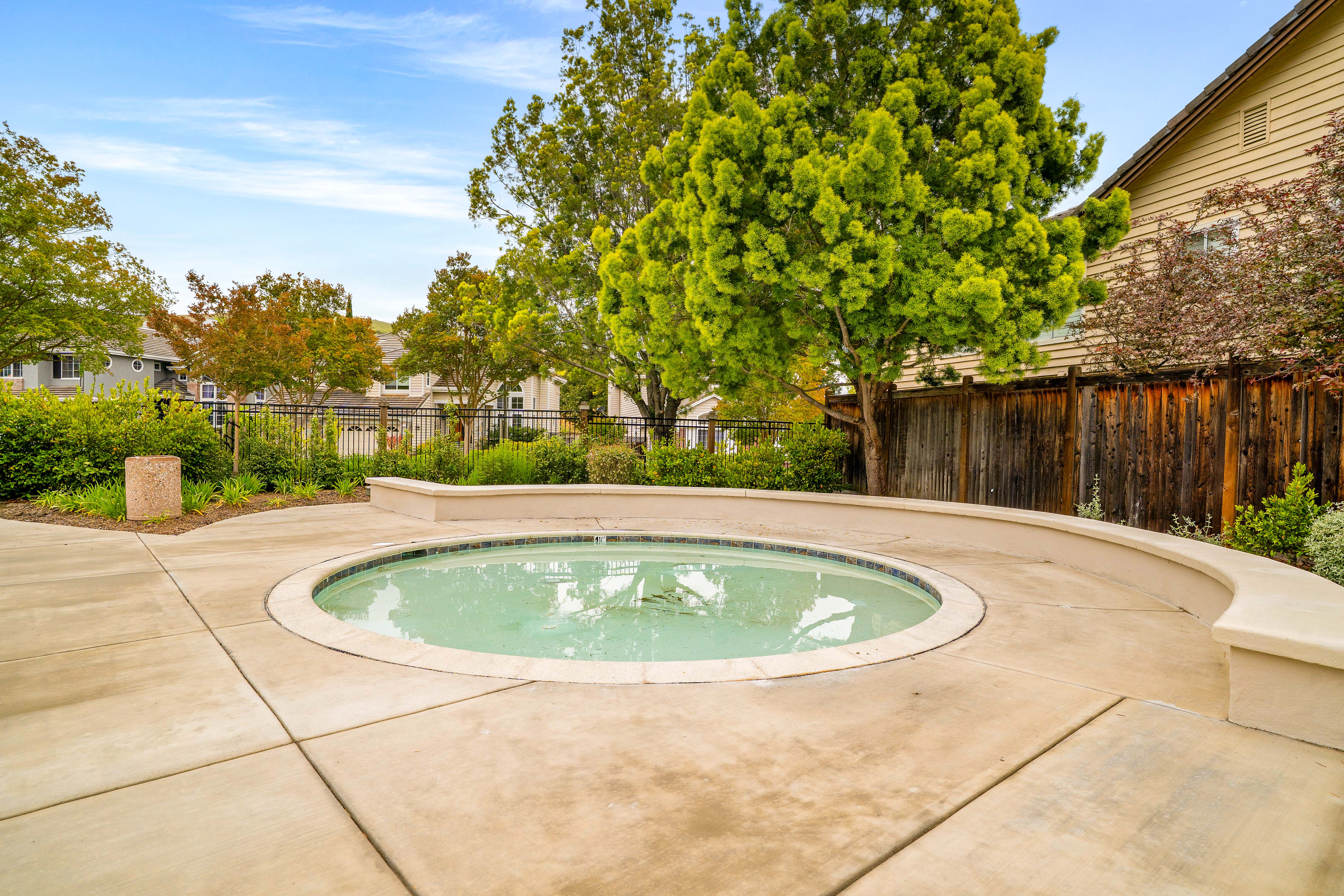This image captures a circular in-ground spa set within a spacious concrete patio area. A curved, low-profile concrete wall provides integrated seating around the perimeter of the spa, which is framed by lush green trees and a wooden privacy fence. The scene is captured from a ground-level perspective, highlighting the outdoor relaxation space in a suburban backyard setting.