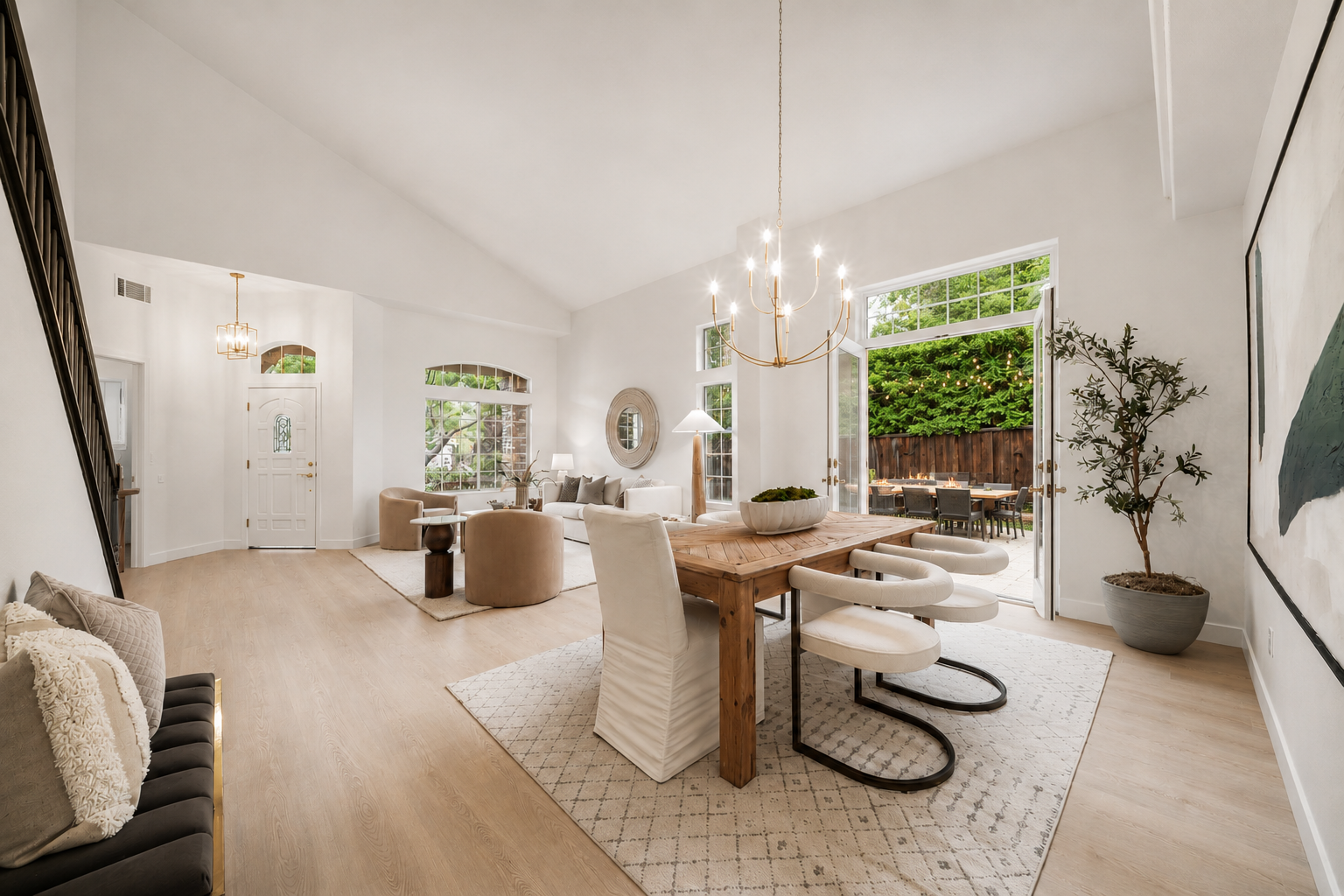 This bright, open-concept dining area flows seamlessly into a living space, featuring light wood flooring and a vaulted ceiling. A rustic wooden dining table is surrounded by modern, curved-back chairs under a gold-toned chandelier, while large glass doors open to an outdoor patio. The neutral color palette and minimalist decor create an airy, sophisticated atmosphere perfect for entertaining.
