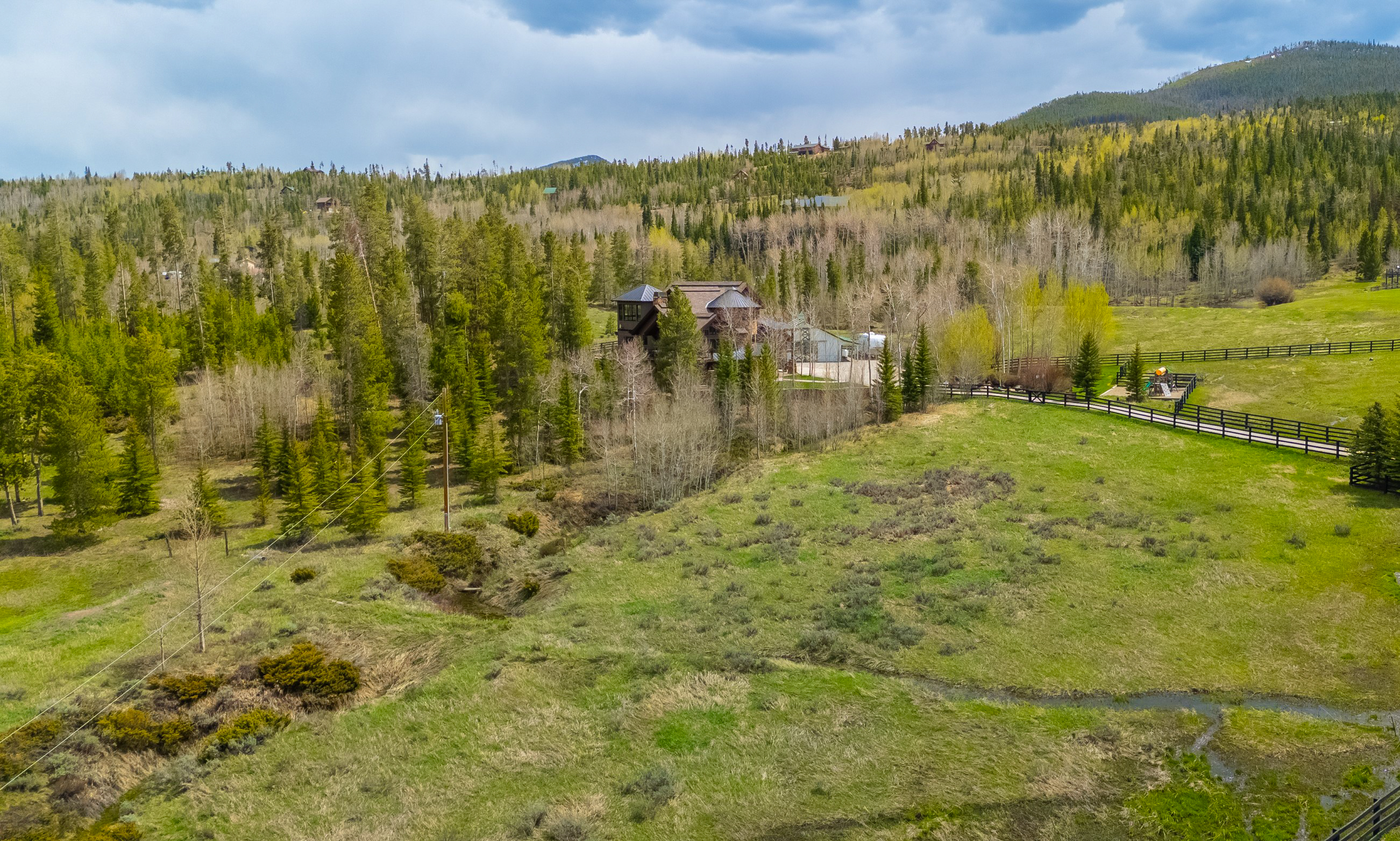 This aerial view captures a large, multi-story mountain home nestled within a dense forest of pine and aspen trees. The property features expansive green meadows, a winding stream, and a fenced-in area with a small playground, all set against a backdrop of rolling hills under a cloudy sky. The perspective provides a comprehensive look at the home's integration into the natural landscape, highlighting its secluded and serene setting.