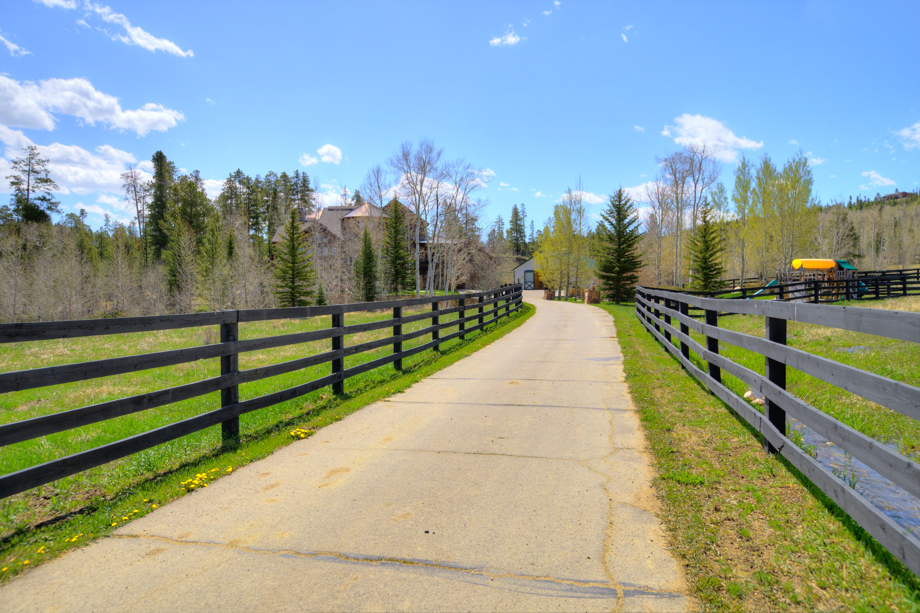 A paved driveway leads through a lush, grassy landscape, framed by dark wooden post-and-rail fencing on both sides. The path curves gently toward a large, multi-story residence nestled among mature pine and aspen trees under a bright blue sky. To the right, a small playground structure is visible, enhancing the property's appeal as a spacious, family-friendly estate.