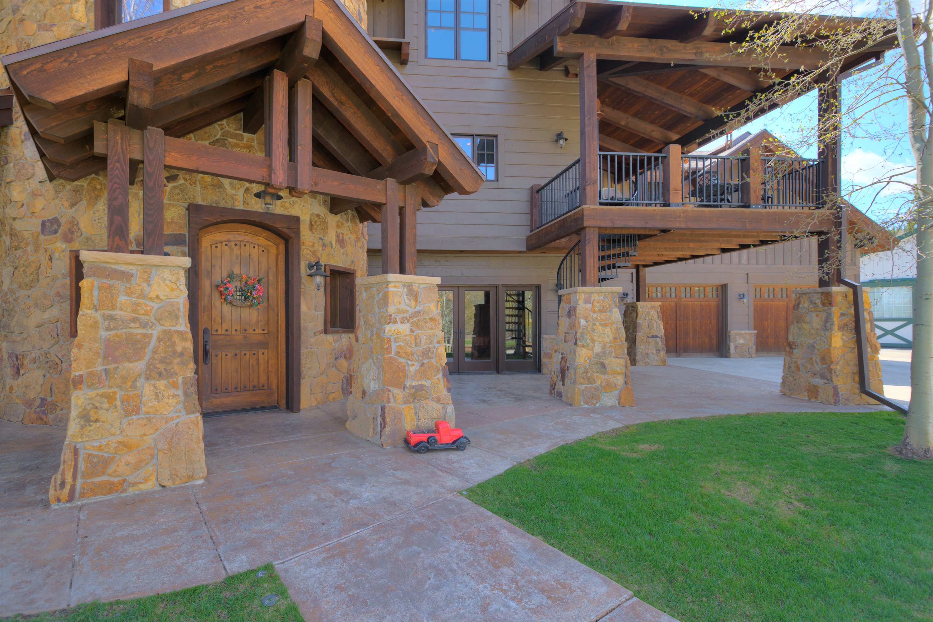 This rustic mountain-style home features a prominent stone-clad entryway with heavy timber framing and a covered porch. The exterior showcases a blend of natural stone masonry and horizontal wood siding, complemented by a large wooden front door and a second-story balcony with a spiral staircase. A small red toy truck sits on the concrete walkway, which leads toward the home's garage area and a lush green lawn.