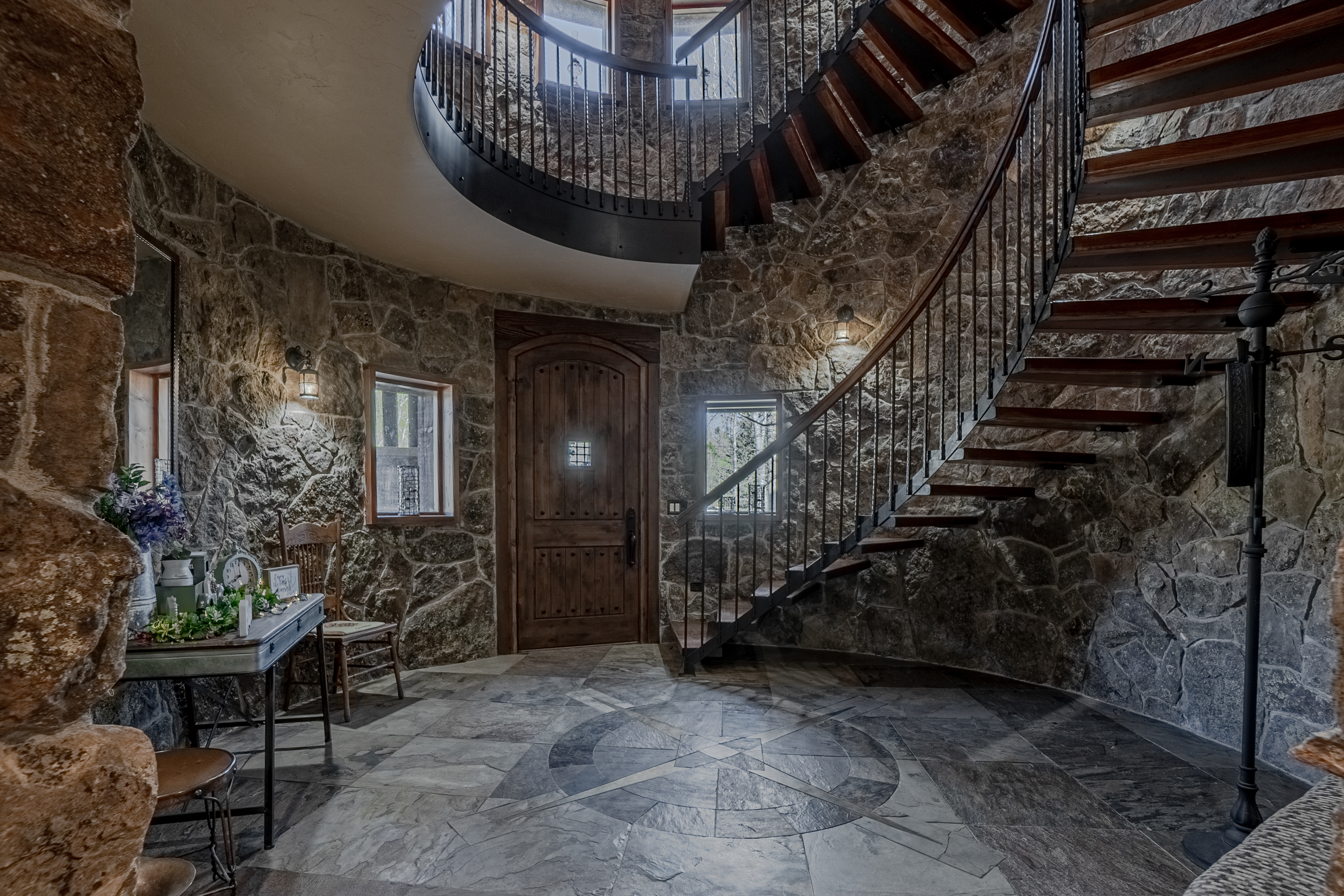 This dramatic entryway features a striking curved staircase with wooden treads and a dark metal railing, set against a backdrop of rustic, textured stone walls. The floor is finished in large, patterned stone tiles arranged in a circular motif, leading toward a heavy, arched wooden front door. The space is illuminated by wall-mounted sconces and natural light from windows flanking the door, creating a grand, castle-like atmosphere.
