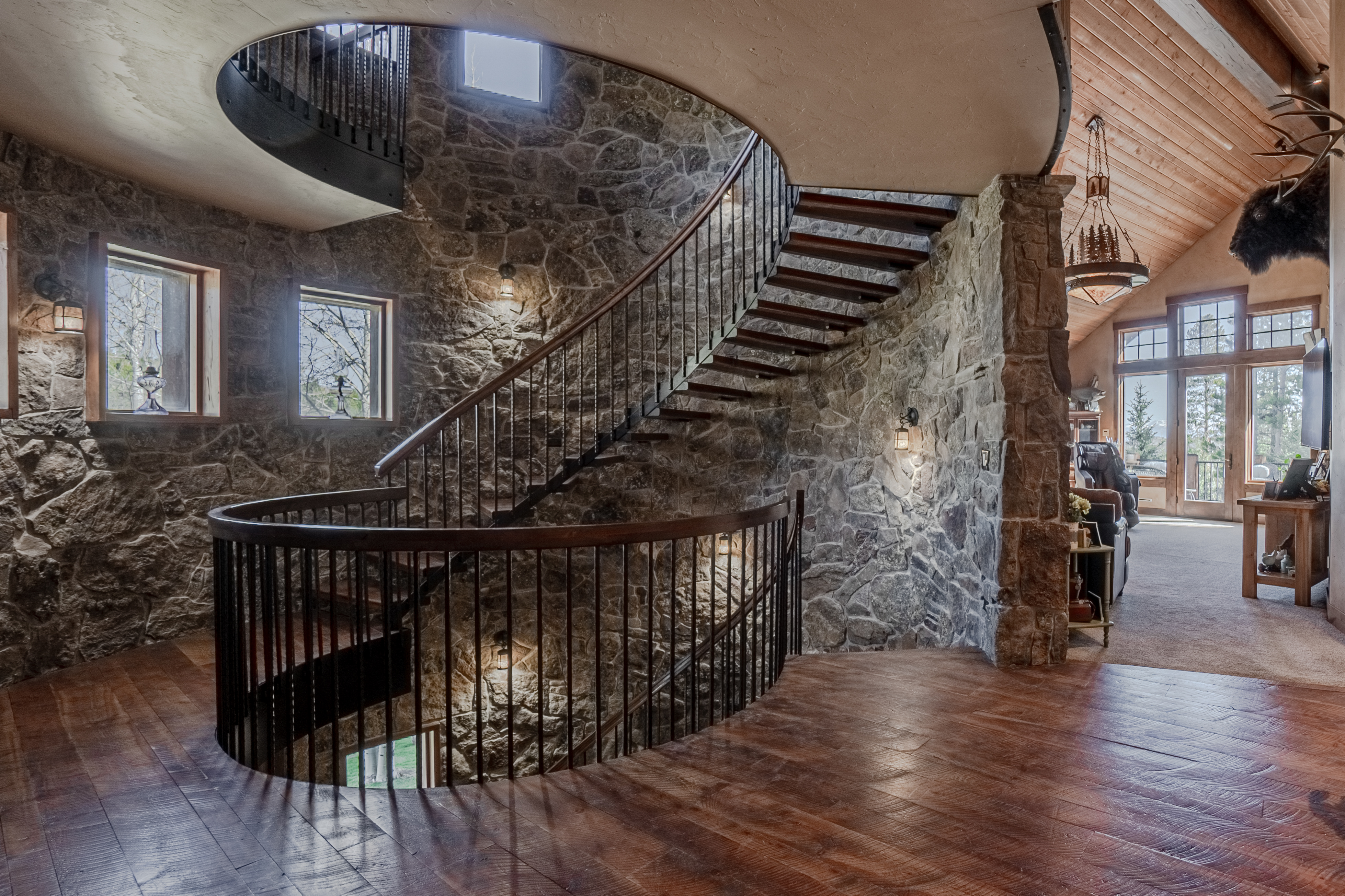 This striking interior space features a prominent curved staircase with dark metal railings and wood treads, set against a dramatic, textured stone wall. The perspective captures the elegant transition between levels, with rich wood flooring and warm lighting creating a rustic, lodge-like atmosphere. Large windows allow natural light to filter in, highlighting the architectural craftsmanship and the open, airy feel of the landing.