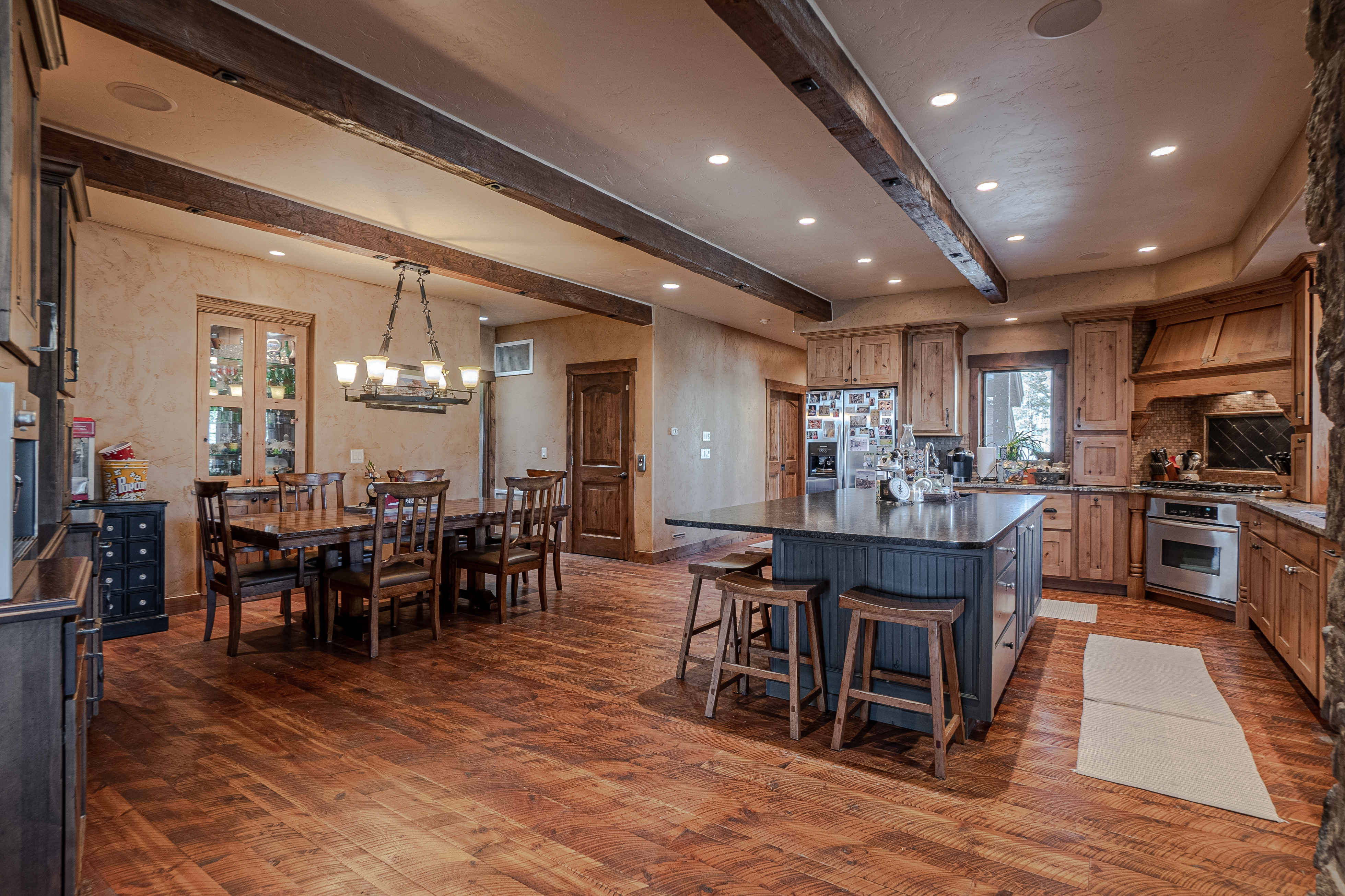 This spacious, open-concept kitchen and dining area features warm wood tones, exposed ceiling beams, and rustic textured walls. A large central island with a dark base and stone countertop provides ample seating, while the adjacent dining space is anchored by a traditional chandelier. The room is finished with rich wood flooring and high-end stainless steel appliances, creating a cozy yet sophisticated mountain-lodge aesthetic.