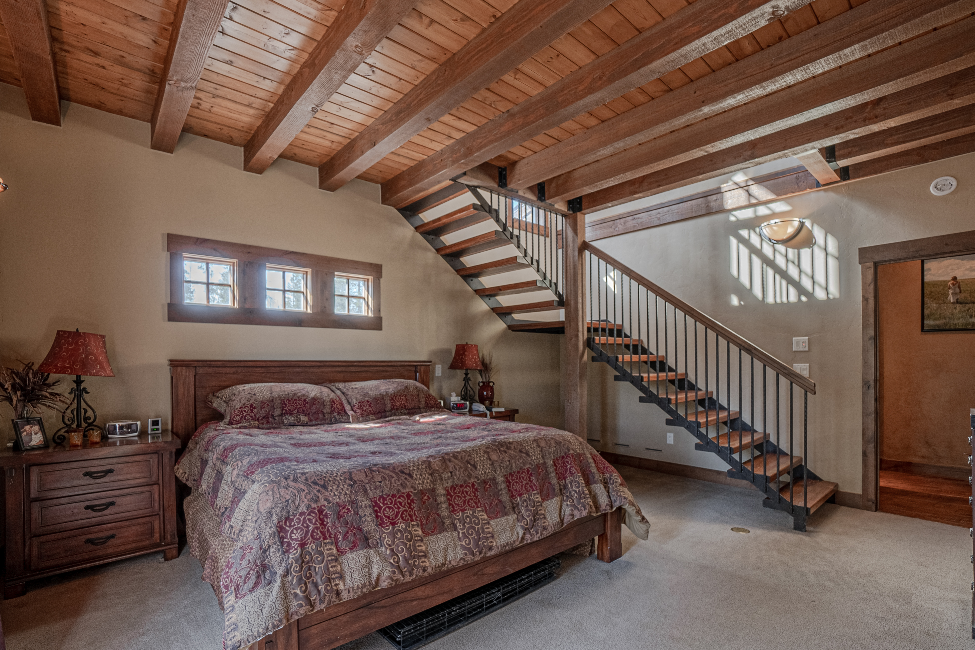 This rustic primary bedroom features a prominent wooden bed frame with a patterned quilt and exposed timber ceiling beams. A striking open-tread staircase with a metal railing ascends from the room, creating a unique architectural focal point. The space is finished with neutral walls, carpeted flooring, and a set of three small windows above the headboard, offering a warm and cozy atmosphere.