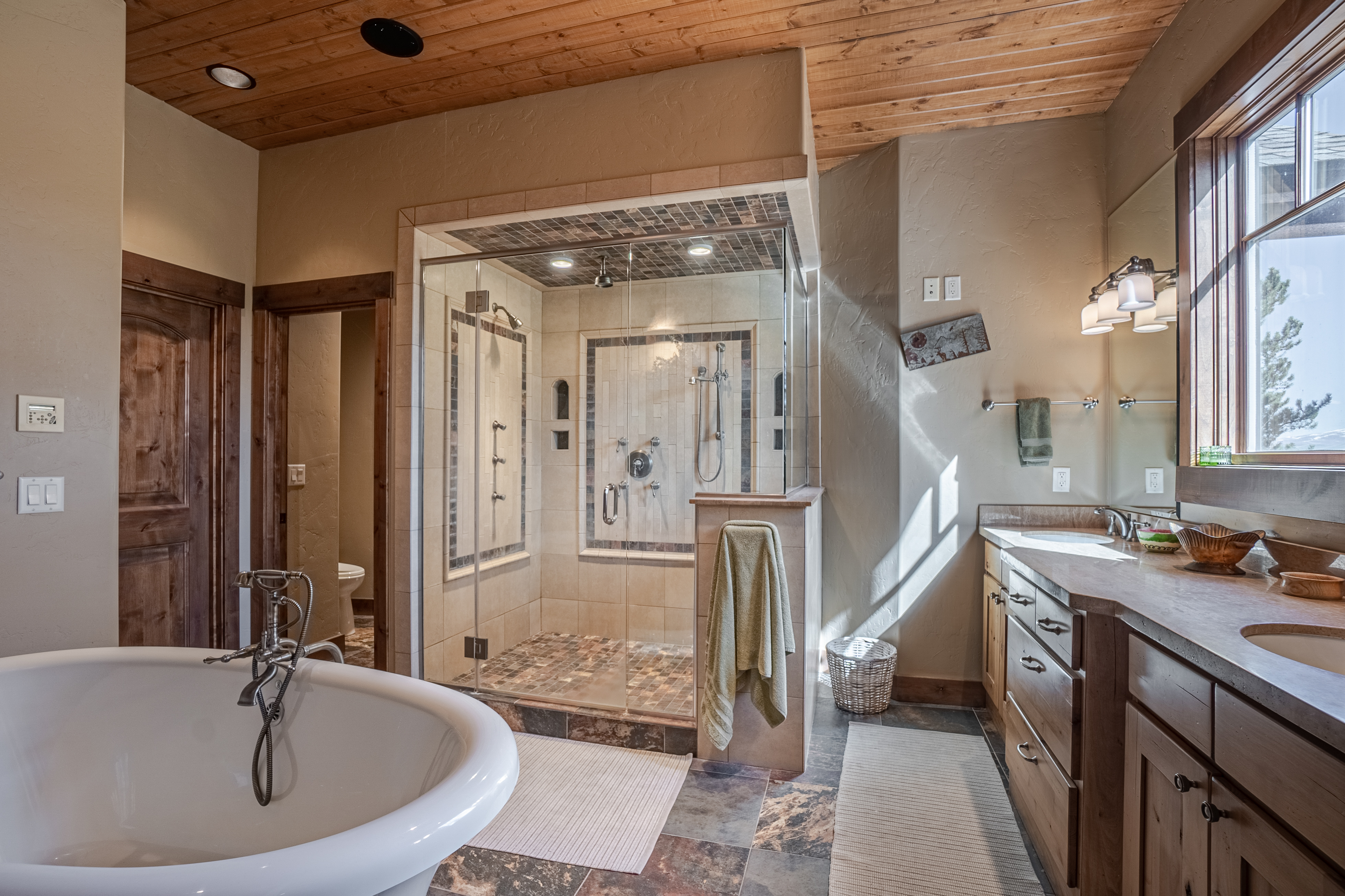 This spacious primary bathroom features a rustic aesthetic with a wood-plank ceiling and warm-toned walls. A large, glass-enclosed walk-in shower with stone tile accents sits centrally, flanked by a freestanding soaking tub in the foreground and a dual-sink vanity with stone countertops to the right. The room is bathed in natural light from a large window, creating an inviting and luxurious atmosphere.