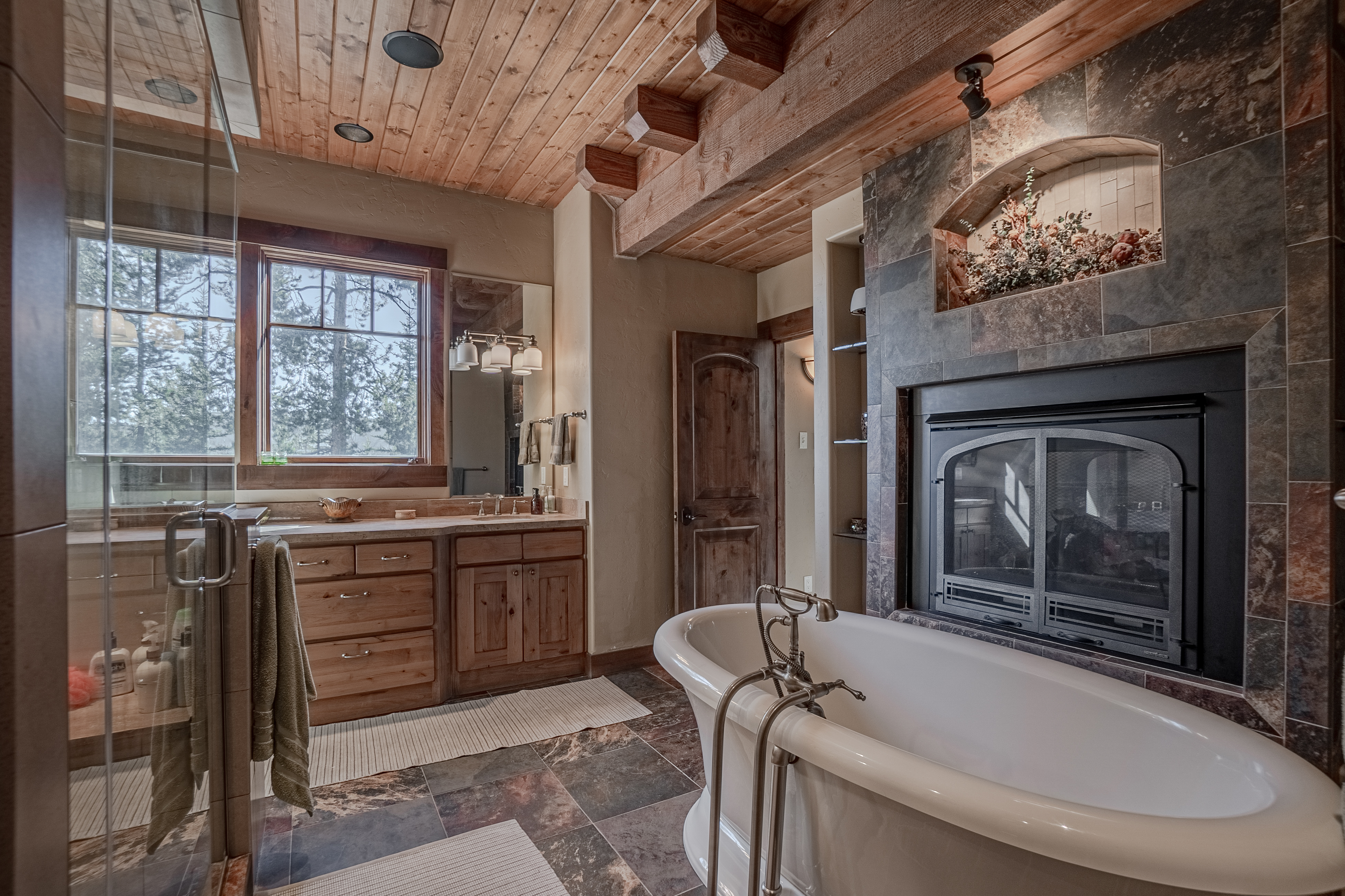 This rustic primary bathroom features a freestanding soaking tub positioned in front of a stone-clad chimney breast with an integrated fireplace. The space is accented by exposed wooden ceiling beams, a wood vanity with a stone countertop, and dark patterned tile flooring. A glass-enclosed shower and a window overlooking the trees complete this warm, lodge-inspired retreat.