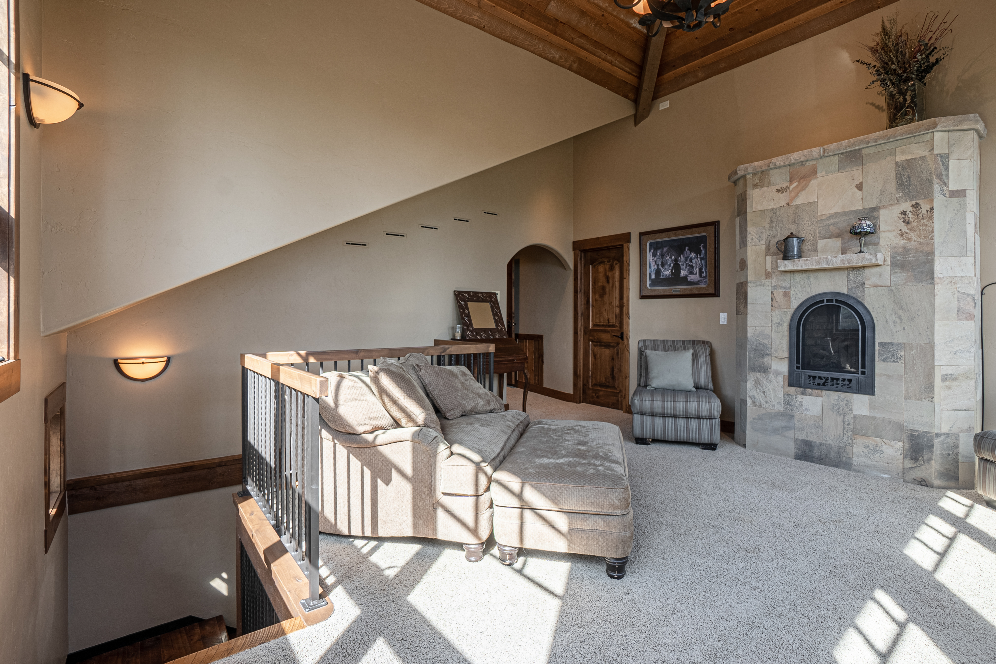 This cozy loft-style living area features a vaulted ceiling with exposed wooden beams and a prominent stone-clad chimney breast housing a fireplace. The space is furnished with a comfortable sofa and ottoman set, complemented by a striped accent chair, all arranged on neutral carpeting. A metal railing overlooks the staircase, while warm wall sconces provide soft lighting, creating an inviting and rustic atmosphere.
