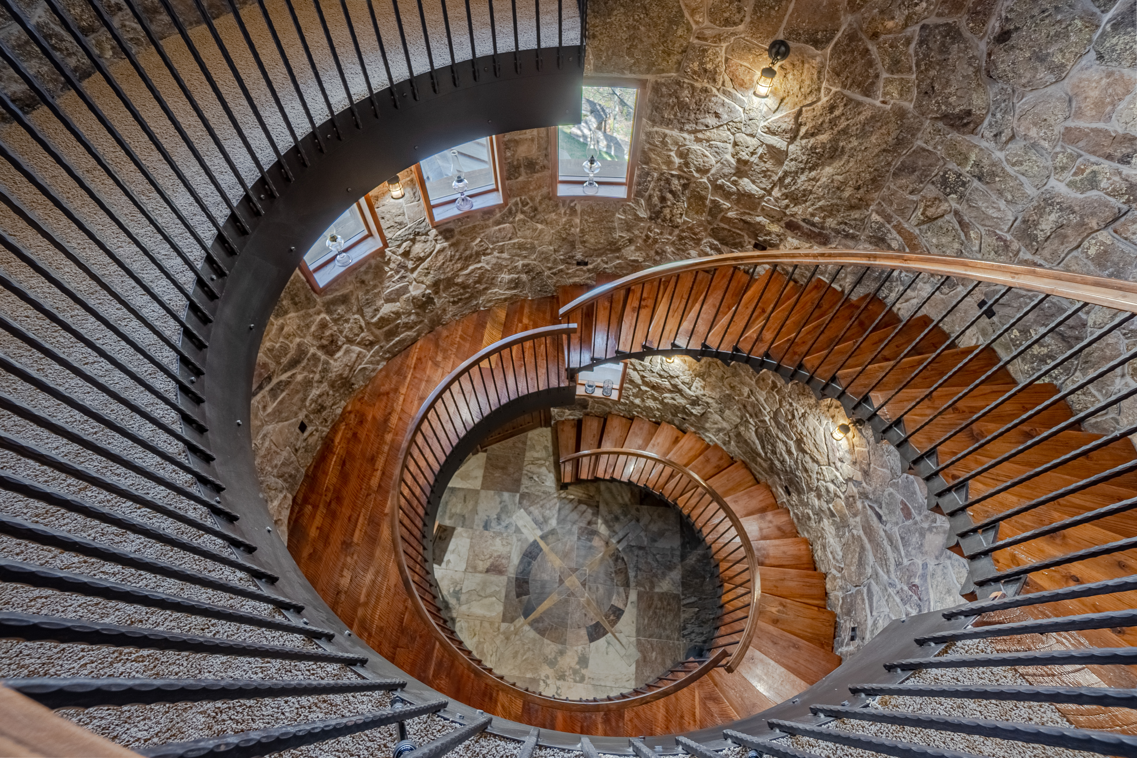 This dramatic high-angle shot captures a grand spiral staircase winding down through a circular stone tower. The staircase features rich wood treads, a dark metal railing, and a stone-clad wall that adds a rustic, architectural character to the space. At the base, a circular floor medallion with a star pattern serves as a focal point, while multiple windows along the curved wall provide natural light to the interior.