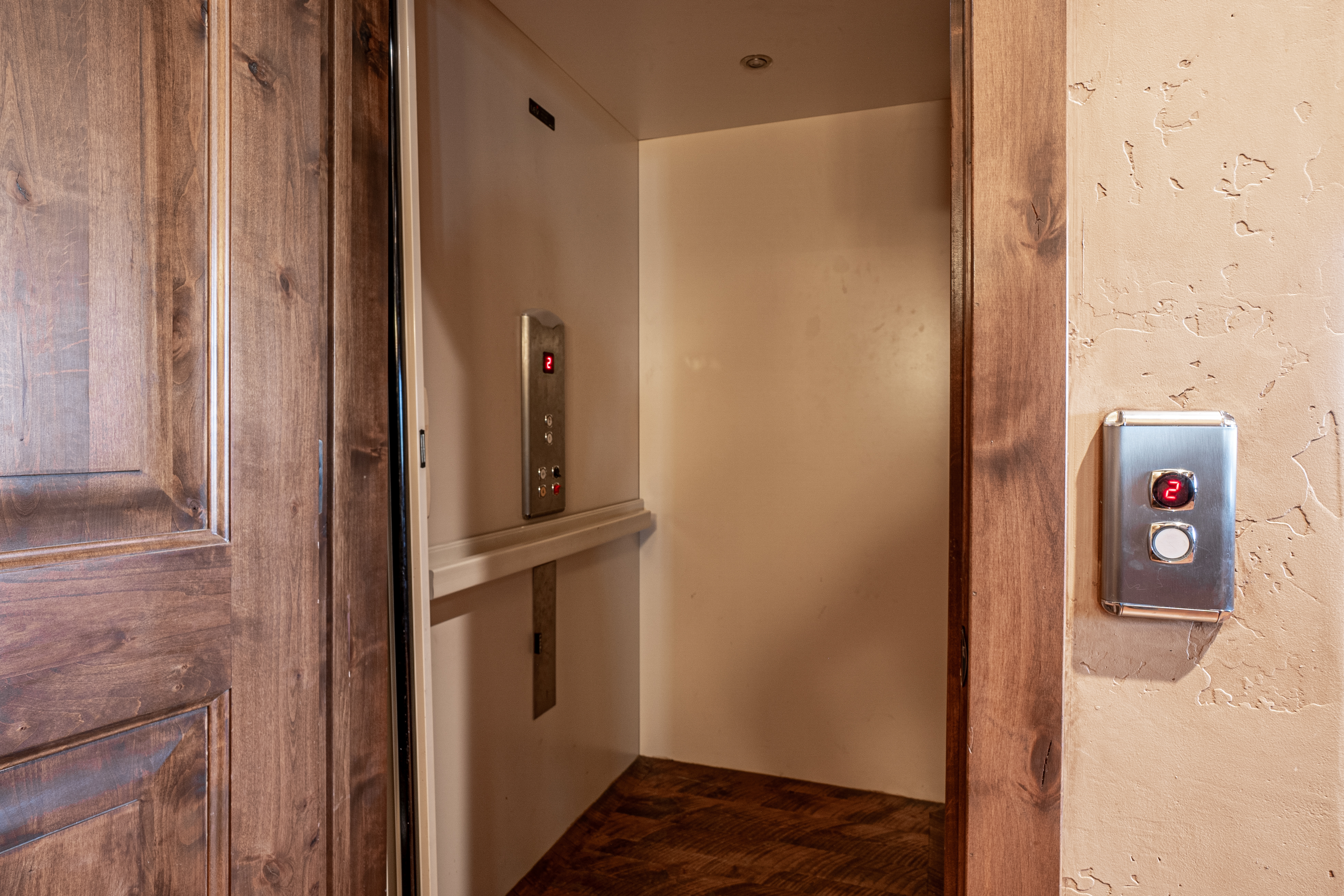 This image captures a residential elevator entrance framed by rich, dark-stained wood trim. The elevator interior features a neutral-toned wall with a control panel, while a matching call button panel is mounted on the textured, light-colored wall to the right. The perspective is centered on the elevator opening, highlighting the convenience and accessibility features of the home.