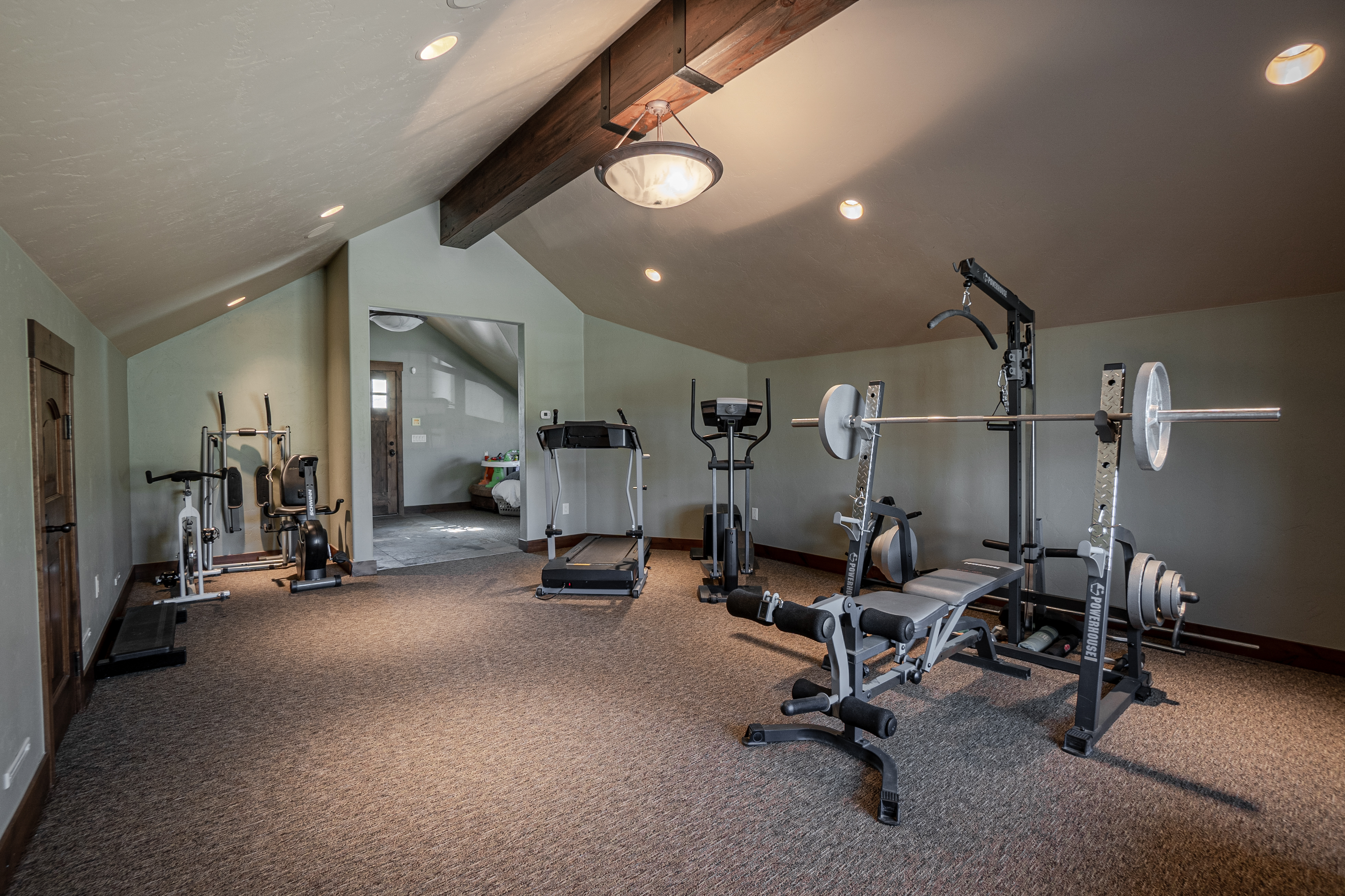 This spacious home gym is located in an attic-style room with vaulted ceilings and a prominent exposed wooden beam. The room is equipped with a variety of fitness equipment, including a treadmill, an elliptical machine, and a weight bench with a barbell rack. The space features neutral-toned walls and wall-to-wall carpeting, creating a functional and dedicated area for exercise.