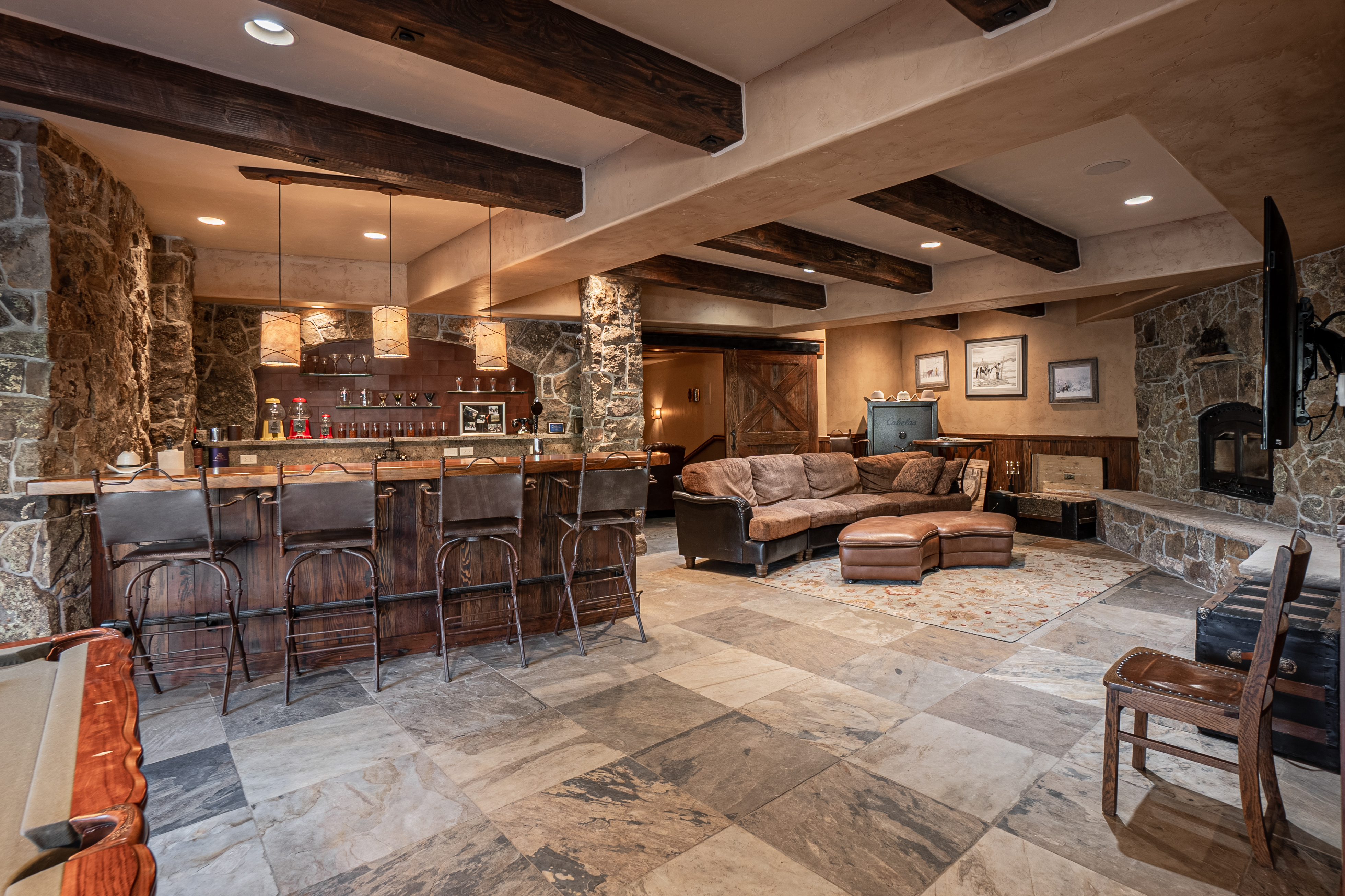 This expansive, rustic-style basement features a large wet bar with a wood-topped counter and four leather bar stools, illuminated by three pendant lights. The living area includes a plush brown sectional sofa, a matching ottoman, and a stone-clad chimney breast housing a fireplace. The space is defined by exposed wooden ceiling beams, stone accent walls, and large stone floor tiles, creating a warm and inviting atmosphere.