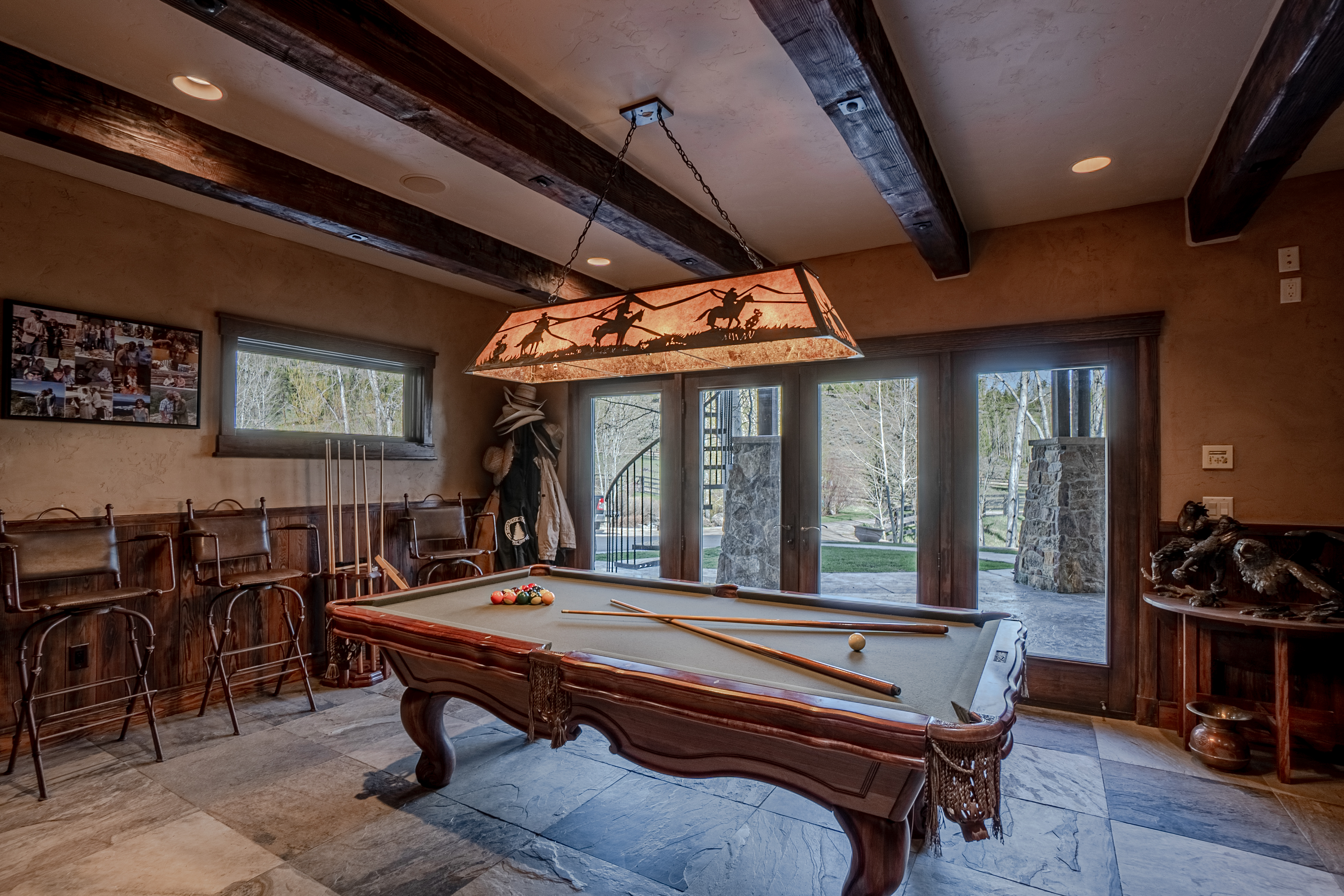 This rustic-style game room features a prominent pool table as the centerpiece, illuminated by a custom western-themed pendant light fixture. The space is characterized by dark wood ceiling beams, stone-tiled flooring, and warm, textured walls, with a set of bar stools and a cue rack arranged along the side. Large glass doors provide a view of the outdoor landscape, creating a seamless transition between the indoor entertainment area and the natural surroundings.