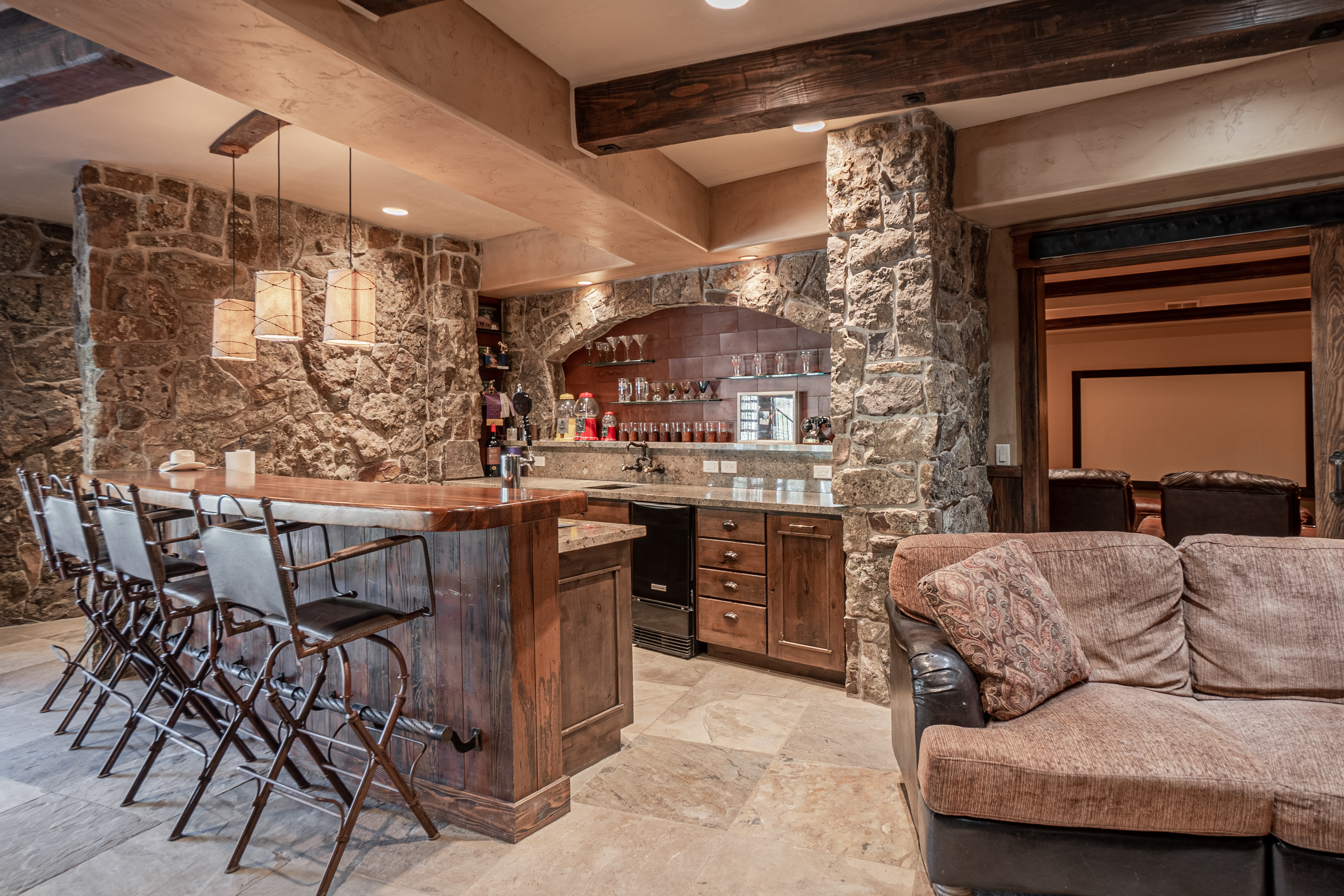 This rustic-style basement bar area features a prominent stone-clad wall and heavy timber ceiling beams, creating a warm and inviting atmosphere. The space includes a wooden bar with high-backed stools, a stone-arched alcove with glass shelving, and a comfortable seating area in the foreground. A doorway in the background leads to a home theater, highlighting the entertainment-focused layout of this lower-level living space.