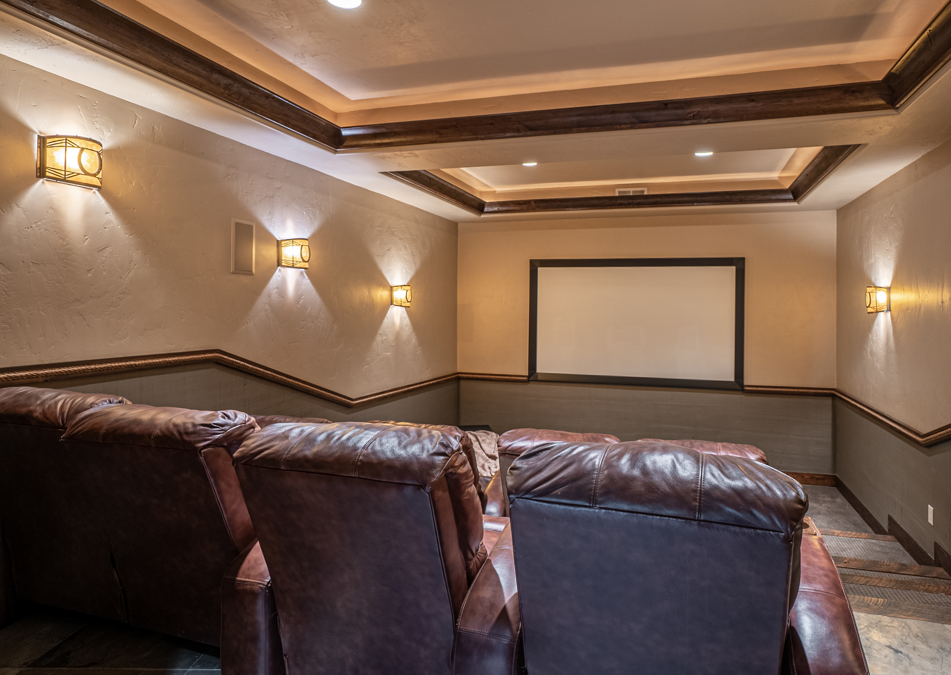 This dedicated home theater room features tiered seating with plush, dark brown leather recliners facing a large projection screen. The space is accented by warm wall sconces, a tray ceiling with recessed lighting, and a two-tone wall treatment with a decorative rope trim. The room offers a cozy, cinematic atmosphere perfect for entertainment.