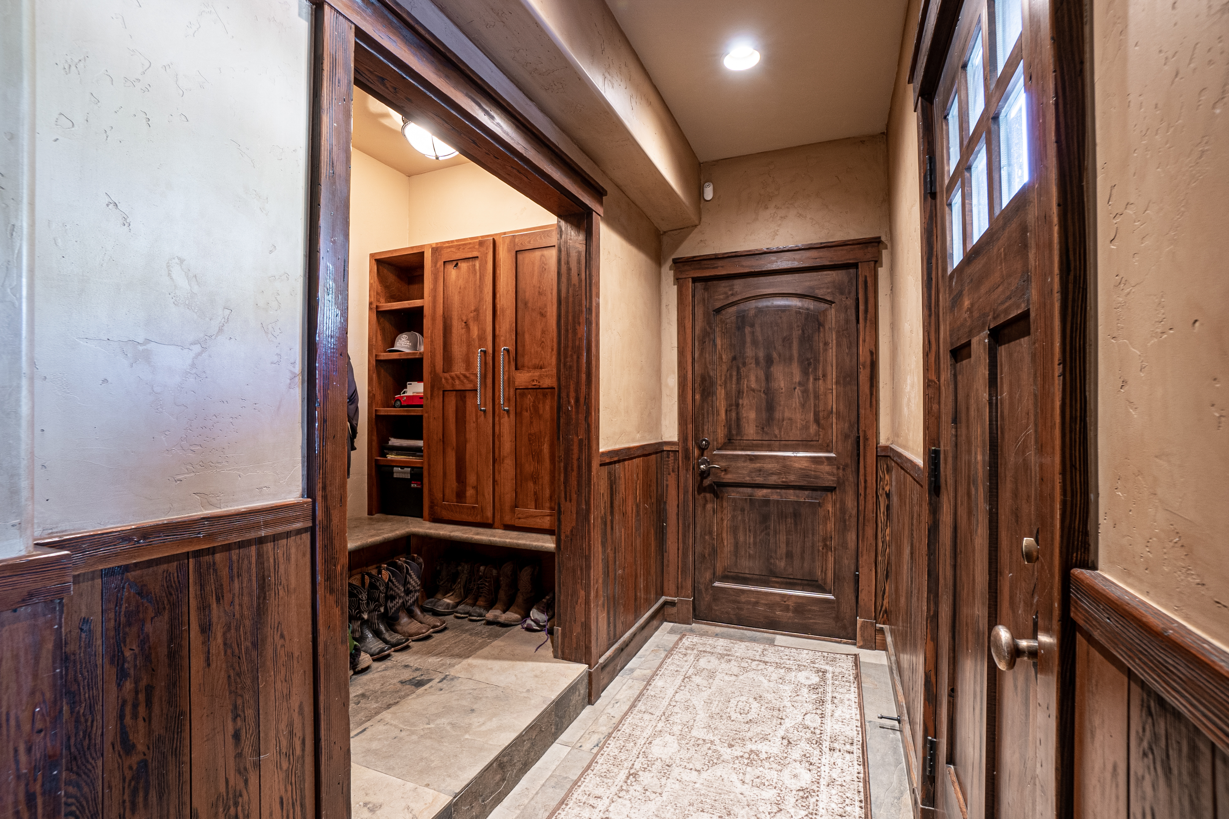 This rustic-style entryway features warm wood paneling on the lower walls and a matching wooden door with glass panes. A recessed mudroom area to the left provides built-in storage cabinets and a bench for footwear, while a patterned runner rug leads the eye down the hallway. The space is illuminated by warm recessed lighting, creating a cozy and inviting atmosphere.