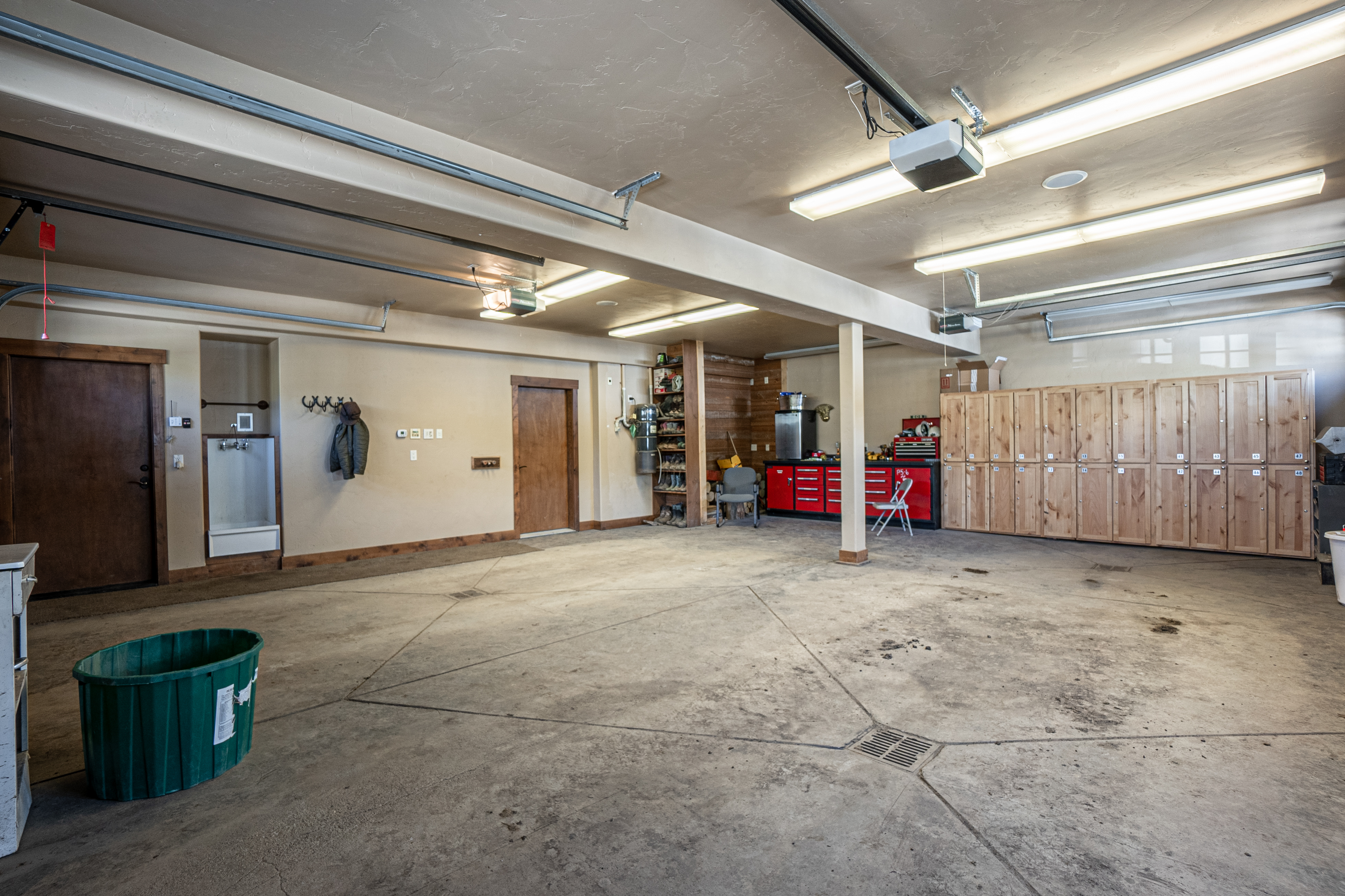 This spacious, multi-car garage features a large concrete floor with integrated drainage and a high, textured ceiling equipped with multiple overhead lighting fixtures. The space is well-organized, offering a dedicated workshop area with red metal cabinets, a wall of light wood storage lockers, and a utility sink station. The perspective is wide-angle, capturing the expansive layout and functional storage solutions that define this versatile utility space.