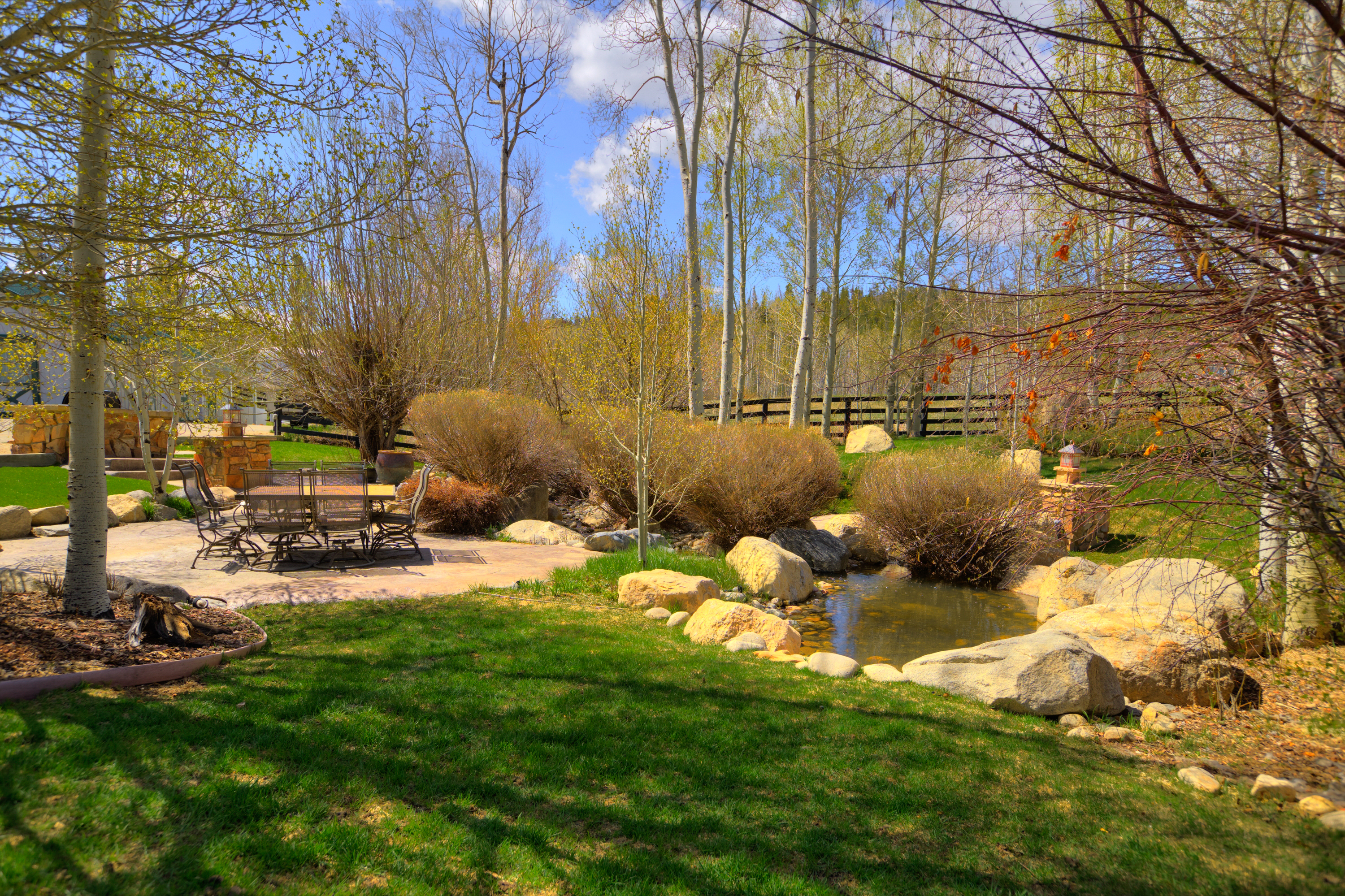 This serene backyard scene features a stone-paved patio area furnished with a dining set, perfect for outdoor entertaining. A naturalistic pond bordered by large boulders and lush landscaping serves as a focal point, set against a backdrop of mature trees and a rustic fence. The perspective is a wide, eye-level shot that captures the peaceful transition from the manicured lawn to the wooded perimeter.