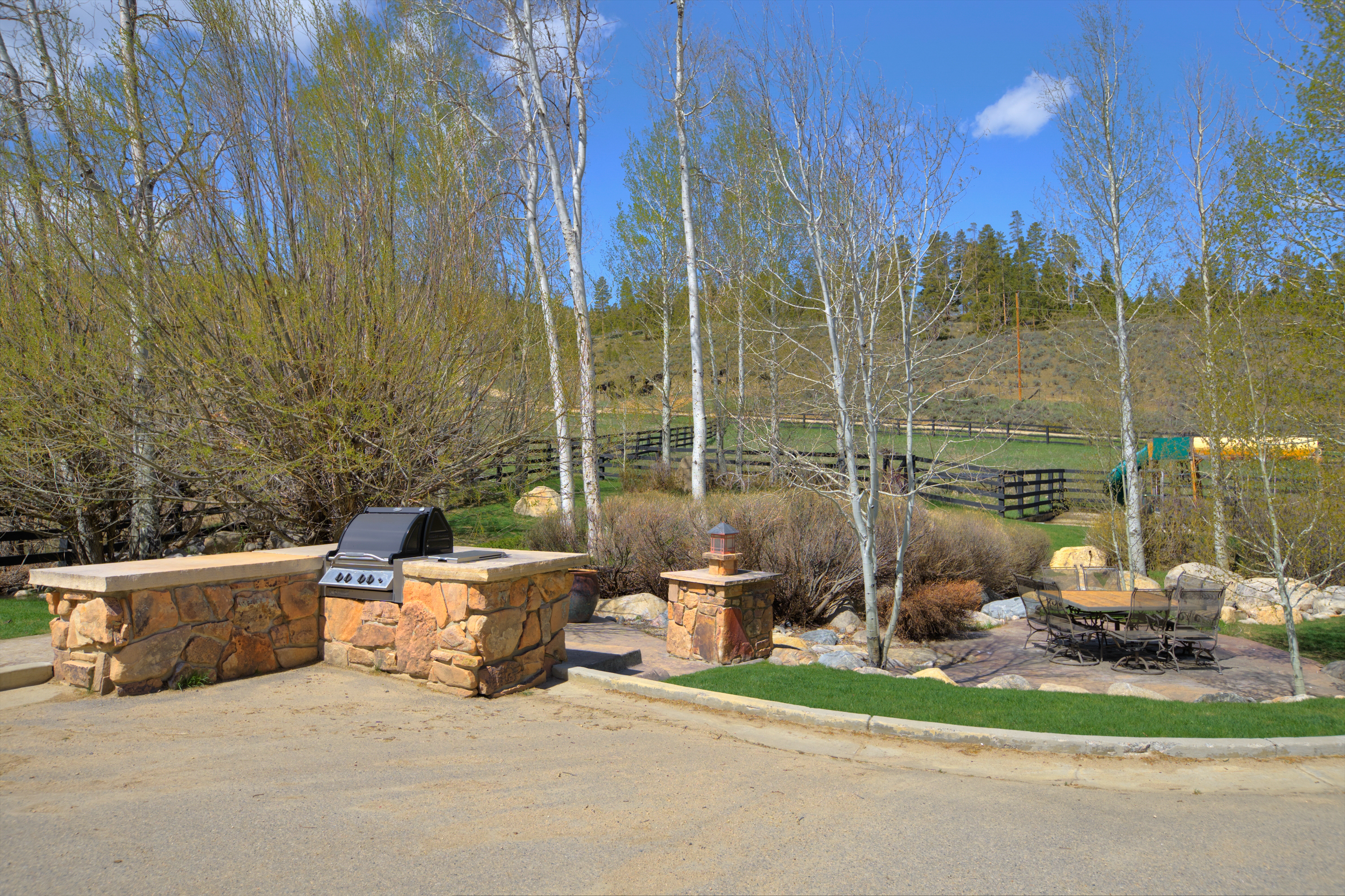 This outdoor living space features a custom stone-clad grill station with a built-in stainless steel barbecue and a matching stone pillar topped with a lantern. A paved patio area provides a dining space with a metal table and chairs, set against a backdrop of mature trees and a fenced pasture. The scene captures a serene, rustic atmosphere perfect for outdoor entertaining in a natural, scenic setting.