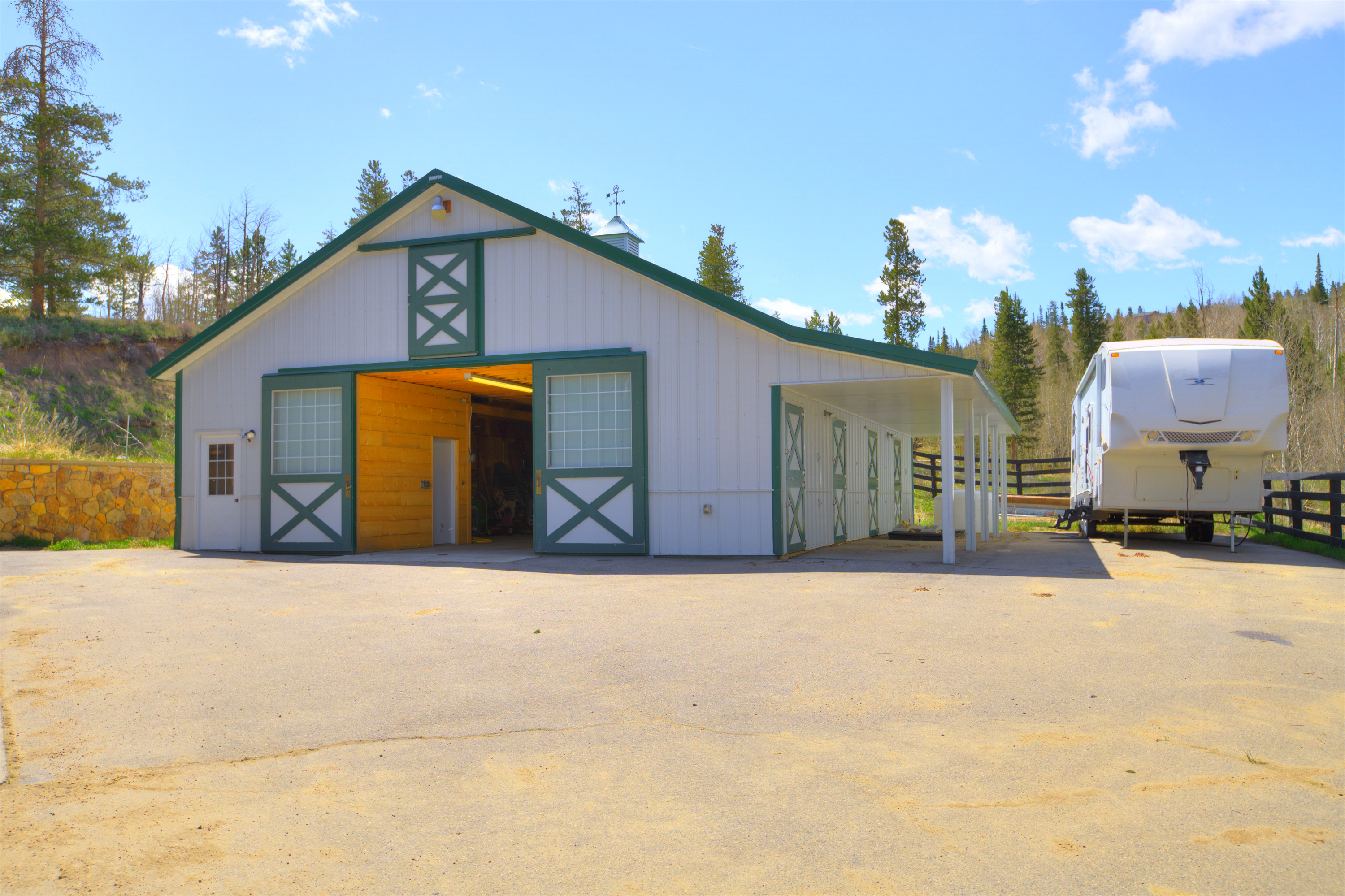 This image showcases a well-maintained, white-sided barn with green trim, featuring large sliding doors that open to reveal a warm, wood-lined interior. To the right, a covered lean-to provides additional storage or shelter, currently housing a large white recreational vehicle. The structure is set against a backdrop of pine trees and a clear blue sky, with a paved driveway leading up to the building.
