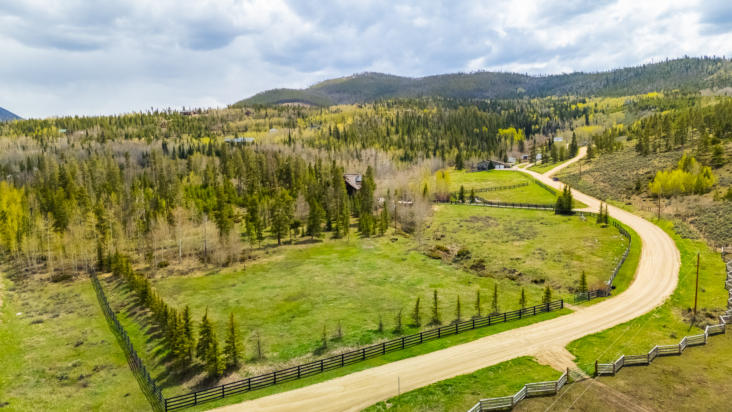 This high-angle aerial perspective captures a sprawling rural property nestled in a mountainous landscape. A winding dirt road traverses the lush green meadow, bordered by classic black post-and-rail fencing that defines the expansive acreage. The scene is framed by dense evergreen forests and rolling hills under a bright, cloudy sky, highlighting the serene and secluded nature of the estate.
