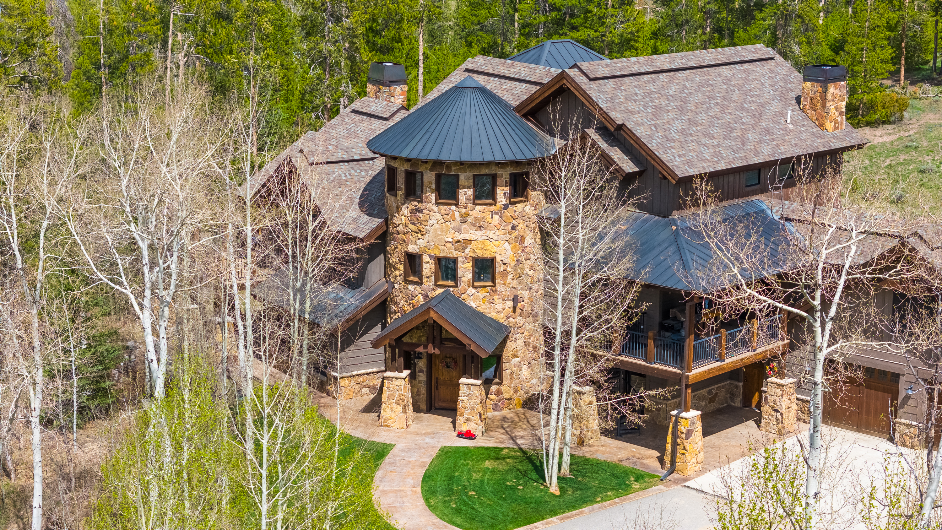 This aerial perspective captures a grand mountain-style residence nestled within a lush, wooded landscape. The home features a prominent stone-clad turret with a dark metal roof, complemented by shingled roofing and warm-toned wood siding. A covered entryway and a spacious elevated deck provide inviting outdoor living spaces, while the surrounding aspen trees and manicured lawn enhance the property's serene, secluded atmosphere.