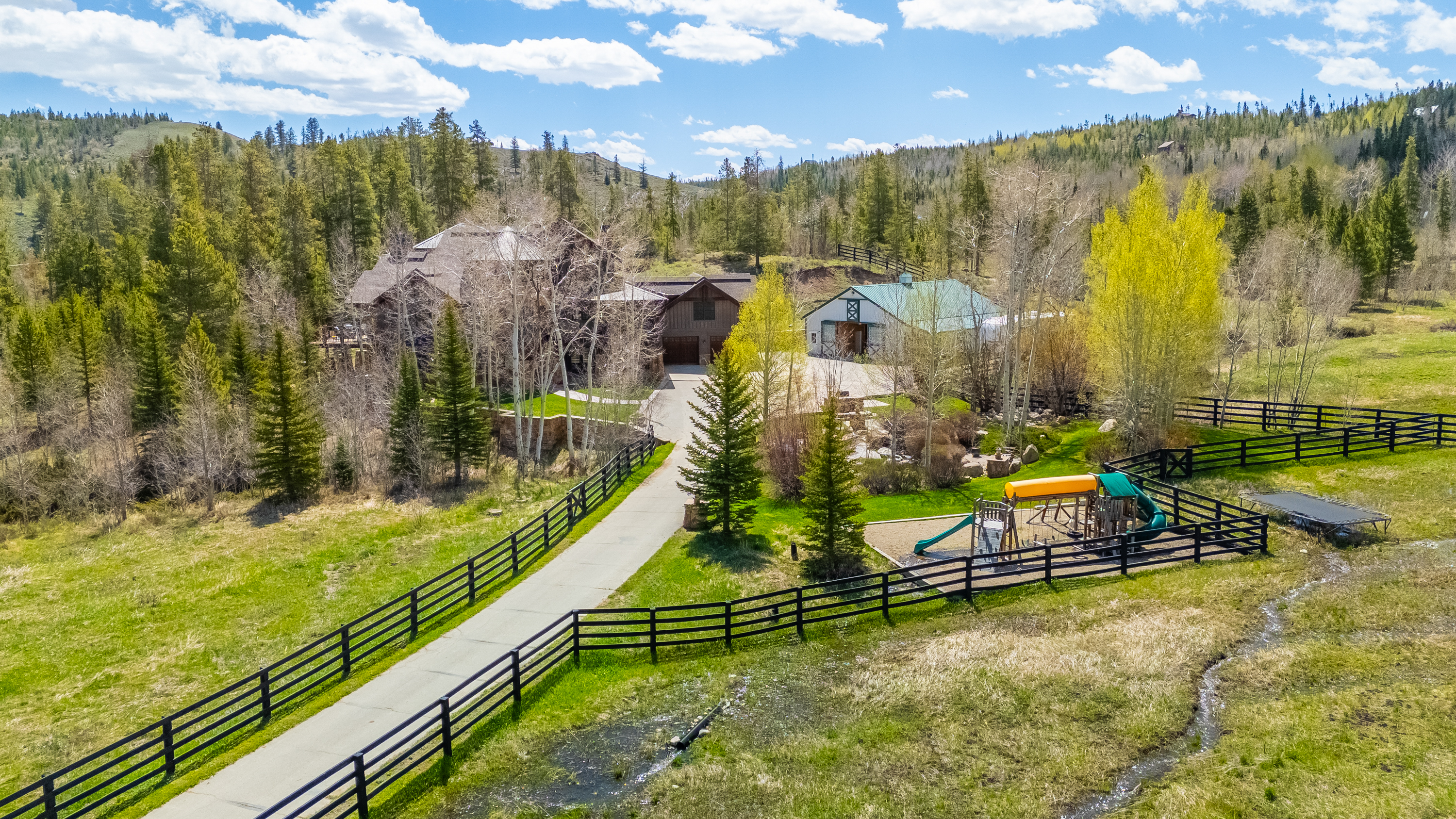 This aerial perspective captures a sprawling mountain estate nestled within a lush, forested landscape. The property features a large, multi-story residence with a rustic aesthetic, an adjacent barn-style structure, and a paved driveway winding through the grounds. A fenced-in play area with a playground set and a small trampoline sits in the foreground, surrounded by open grassy fields and mature evergreen trees.