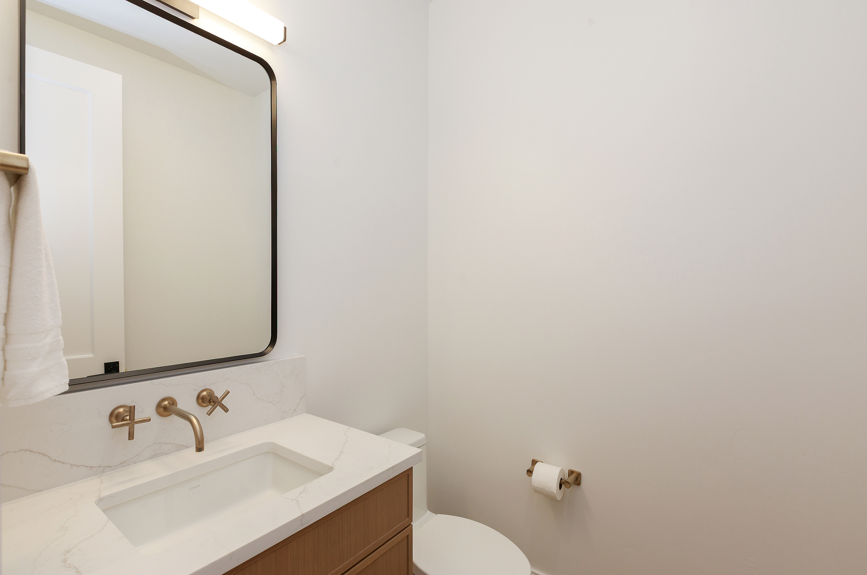 This is a well-lit guest bathroom featuring a modern design. The vanity has a white countertop with a rectangular sink and gold-toned fixtures. A black-framed mirror hangs above the vanity, and a toilet is visible to the right, creating a clean and inviting space.