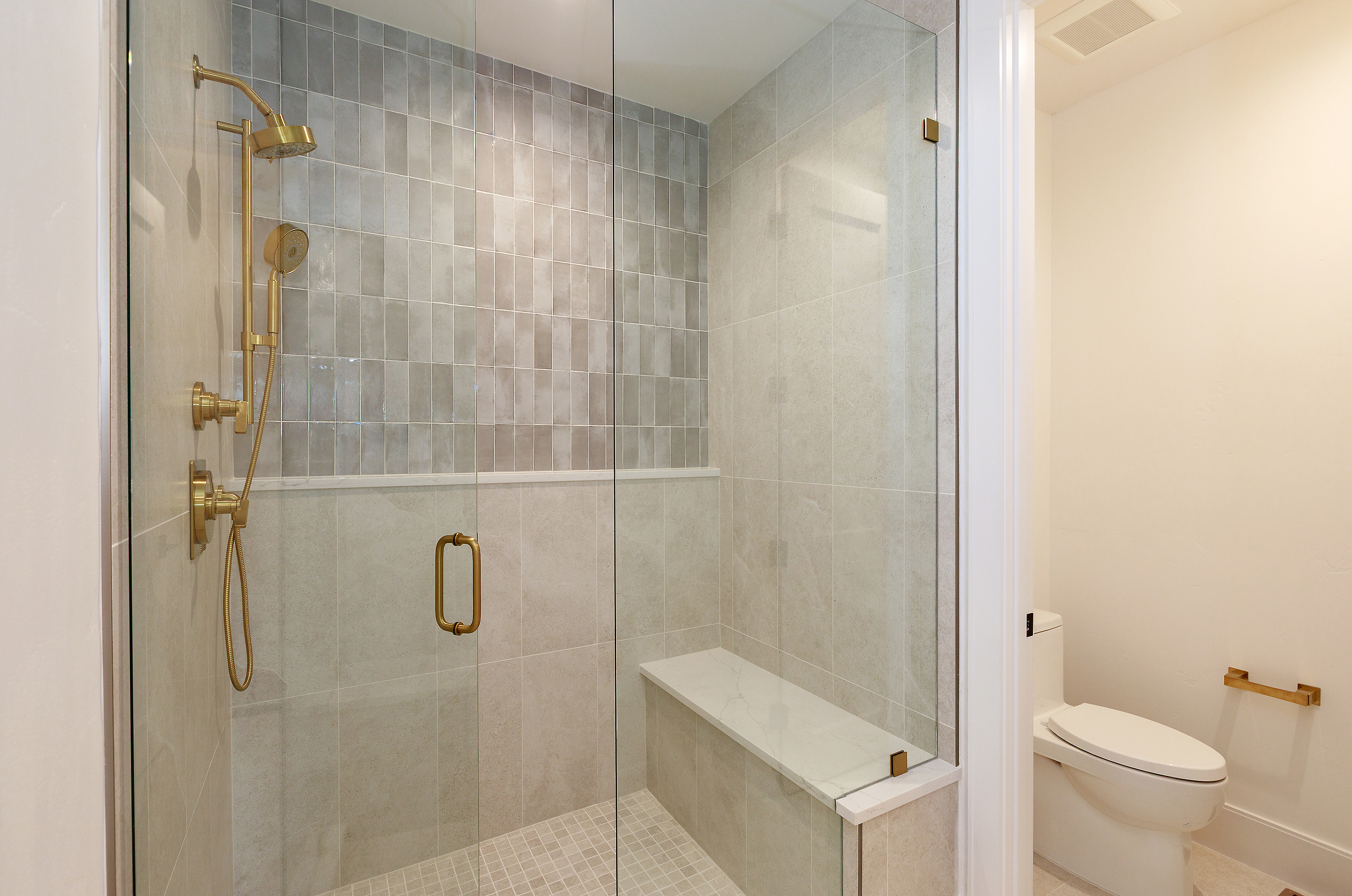 This is a well-lit primary bathroom featuring a modern glass-enclosed shower with gold fixtures and neutral-toned tiling. The shower includes a built-in bench and a handheld shower head. A toilet is visible just outside the shower enclosure, and the overall impression is clean and luxurious.