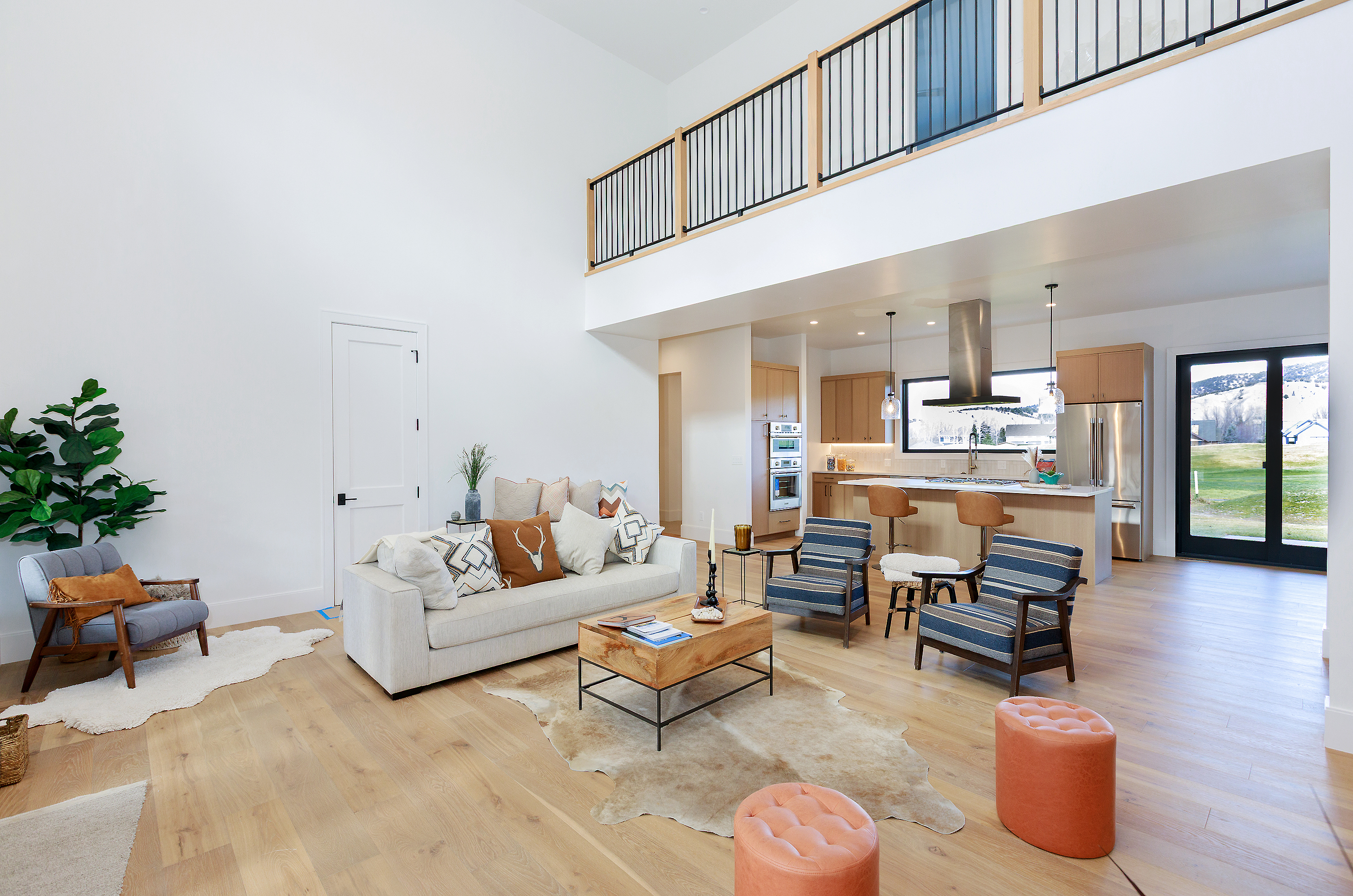This is an interior shot of a modern living room that seamlessly blends into an open-concept kitchen. The living area features a high ceiling, a comfortable sofa with decorative pillows, and stylish armchairs, all arranged around a wooden coffee table on a cowhide rug. The kitchen boasts light wood cabinetry, stainless steel appliances, and a breakfast bar, creating a bright and inviting space.
