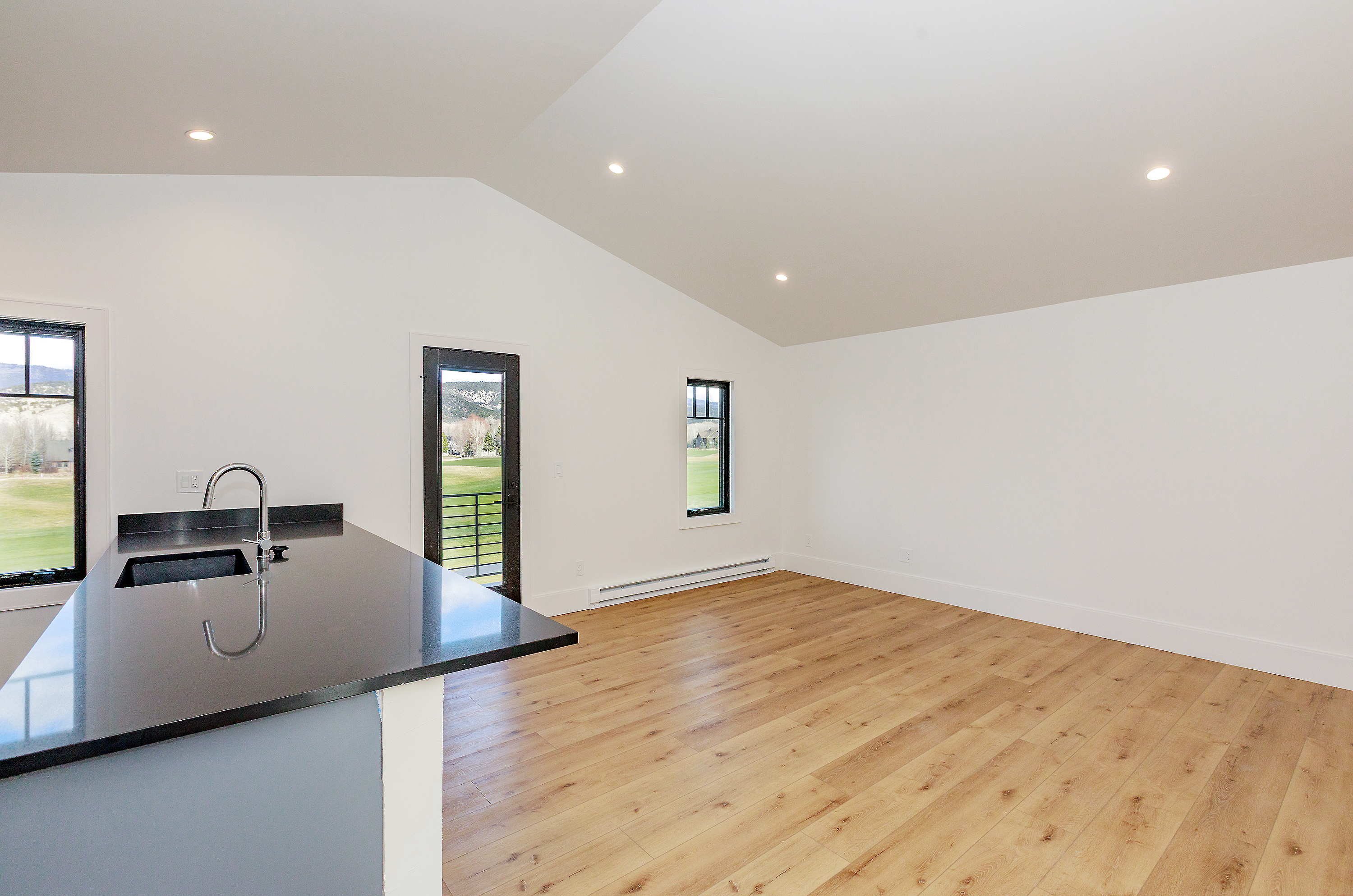 This interior shot showcases a modern kitchen area with sleek, dark countertops and light-colored cabinetry. The space features hardwood flooring and white walls, creating a bright and airy atmosphere. A stainless steel sink and faucet are visible, along with a window offering a view of the outdoors.