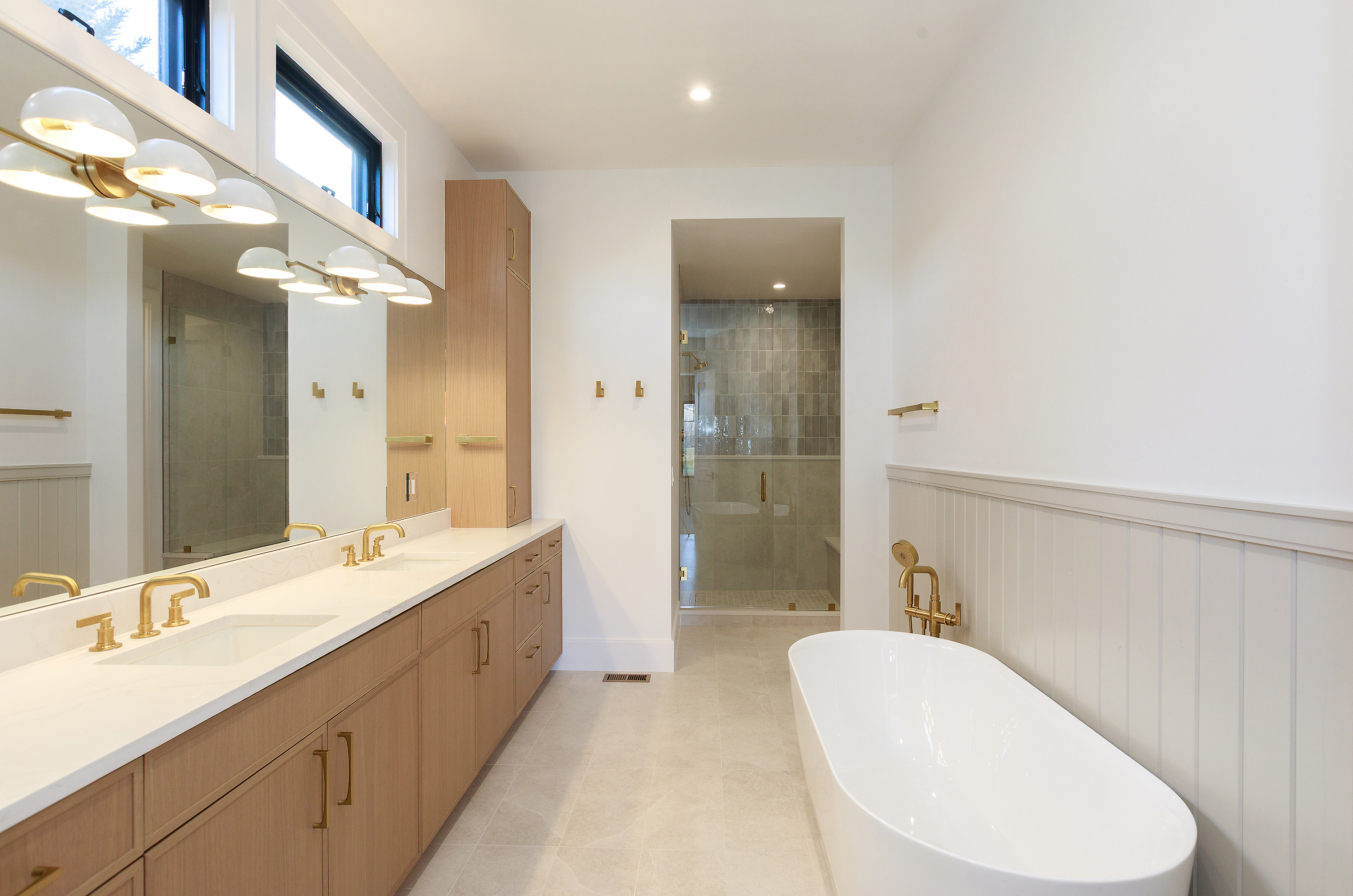 This is a bright and modern primary bathroom featuring a long double vanity with light wood cabinetry, white countertops, and gold fixtures. A freestanding white bathtub sits adjacent to the vanity, complemented by gold hardware and wainscoting. The bathroom also includes a glass-enclosed shower, creating a luxurious and spa-like atmosphere.