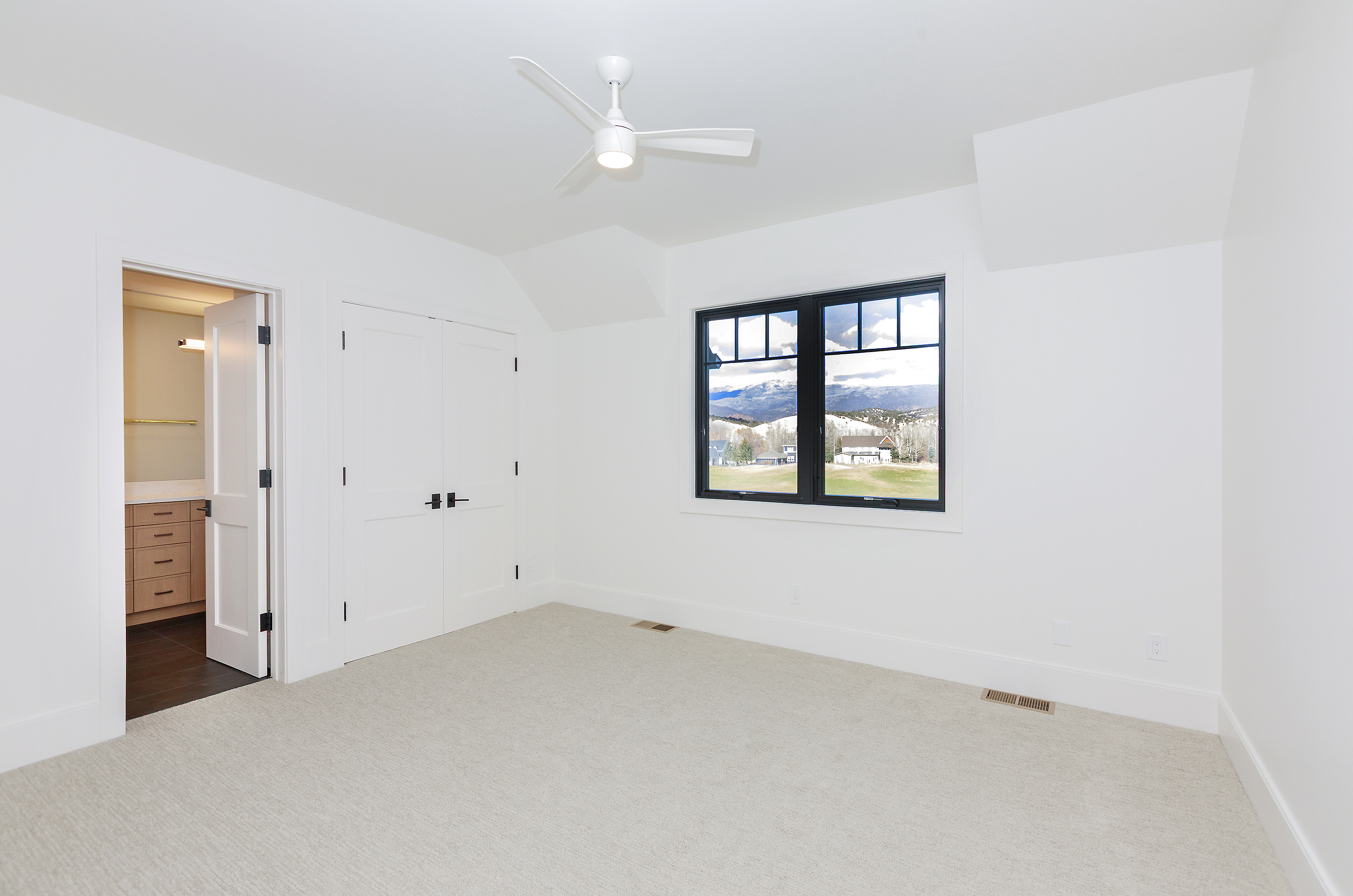 This is a bright and airy bedroom featuring white walls, trim, and a light-colored carpet. A black-framed window offers a view of the landscape outside. The room includes a ceiling fan, closet, and an open doorway to a bathroom or dressing area, creating a clean and modern aesthetic.