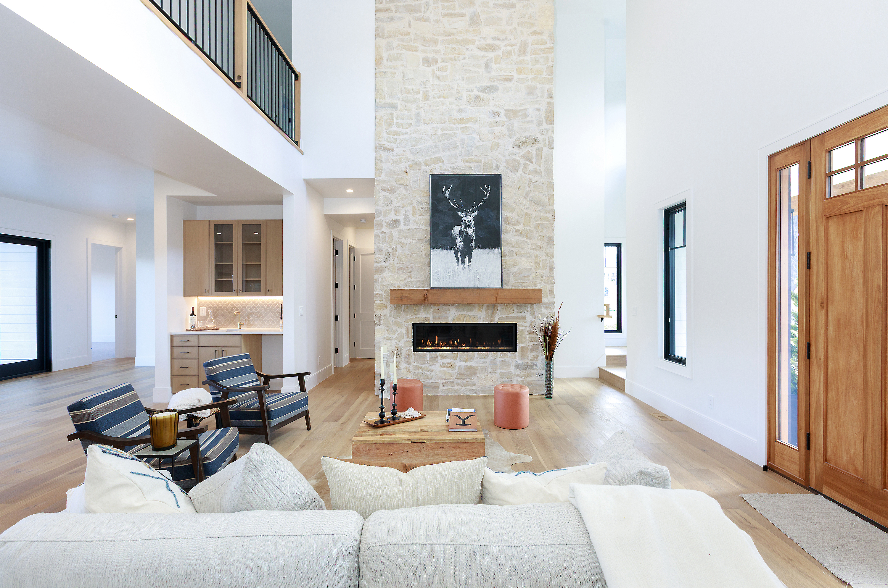 This is an interior shot of a modern living room featuring a stone fireplace with a mounted deer painting. The room is spacious with high ceilings and light hardwood floors. The furniture includes a light-colored sofa, striped armchairs, and a wooden coffee table, creating a comfortable and stylish living space.