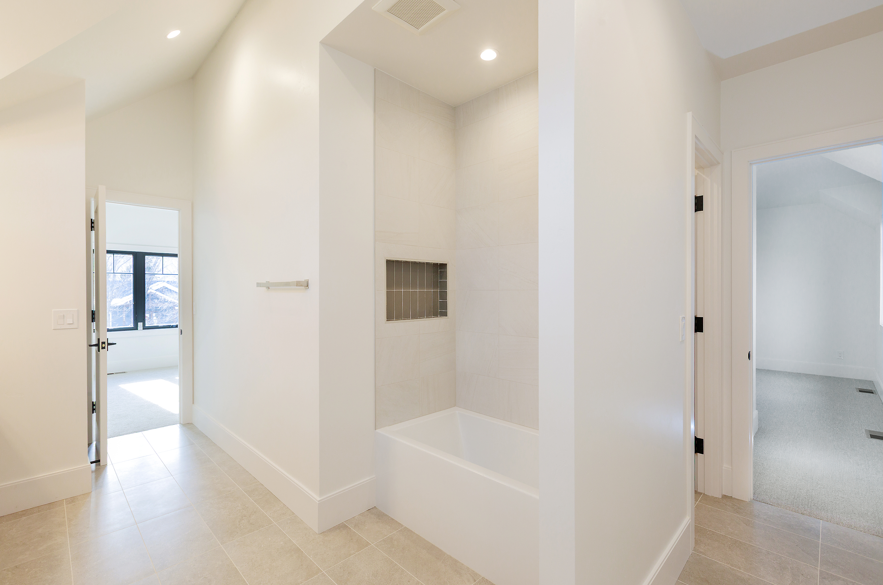This is a bright and clean bathroom featuring a built-in bathtub with tiled walls and a niche for storage. The walls are painted white, and the floor is tiled in a neutral tone. The bathroom has two doorways, one leading to another room and the other to a bedroom, creating a sense of spaciousness and flow.