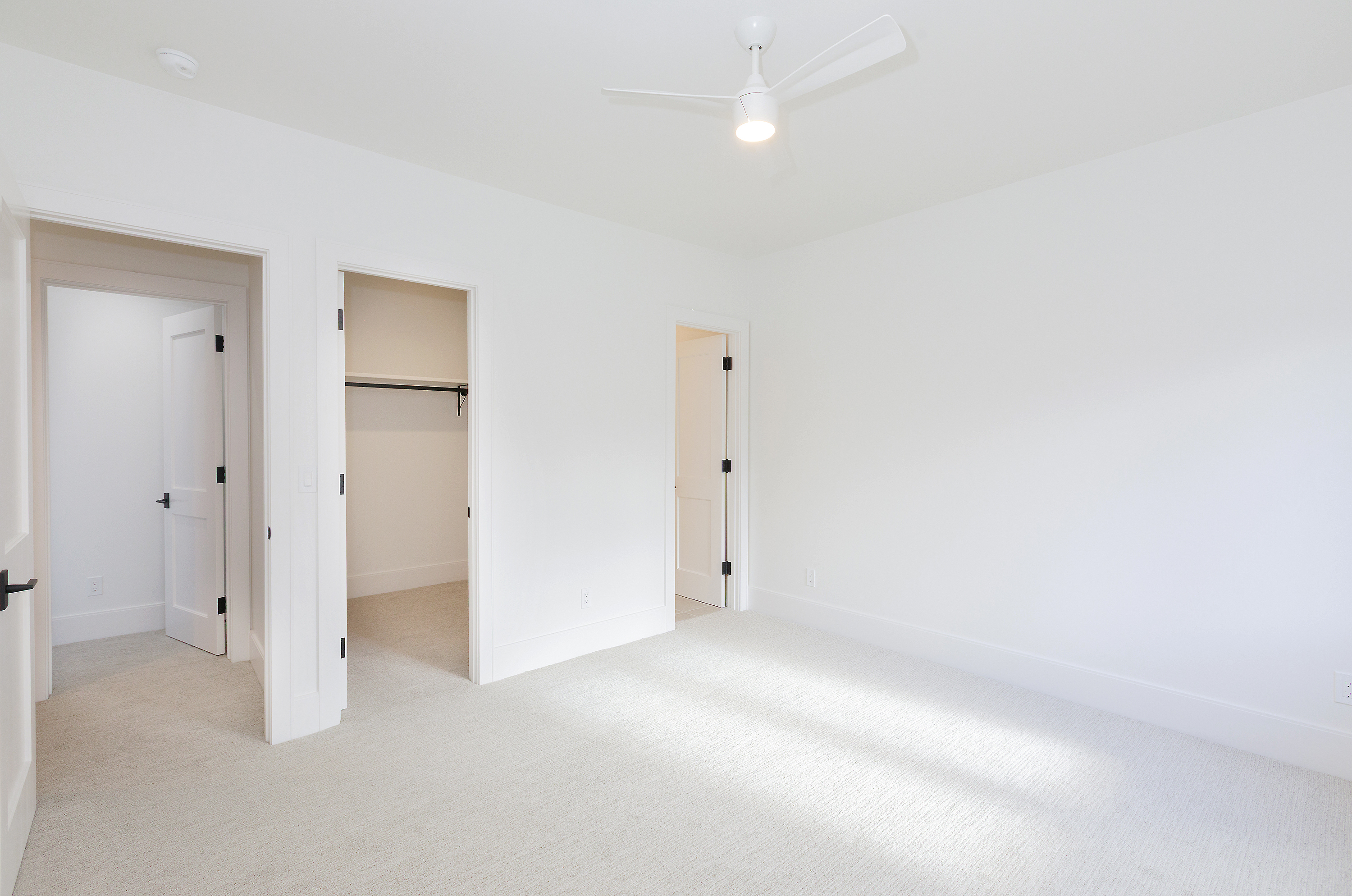 This is a bright, minimalist bedroom with white walls, carpet, and trim. The room features a ceiling fan with a light and three doorways, one leading to a closet. The overall impression is clean and spacious, ideal for a guest bedroom or secondary bedroom in a modern home.