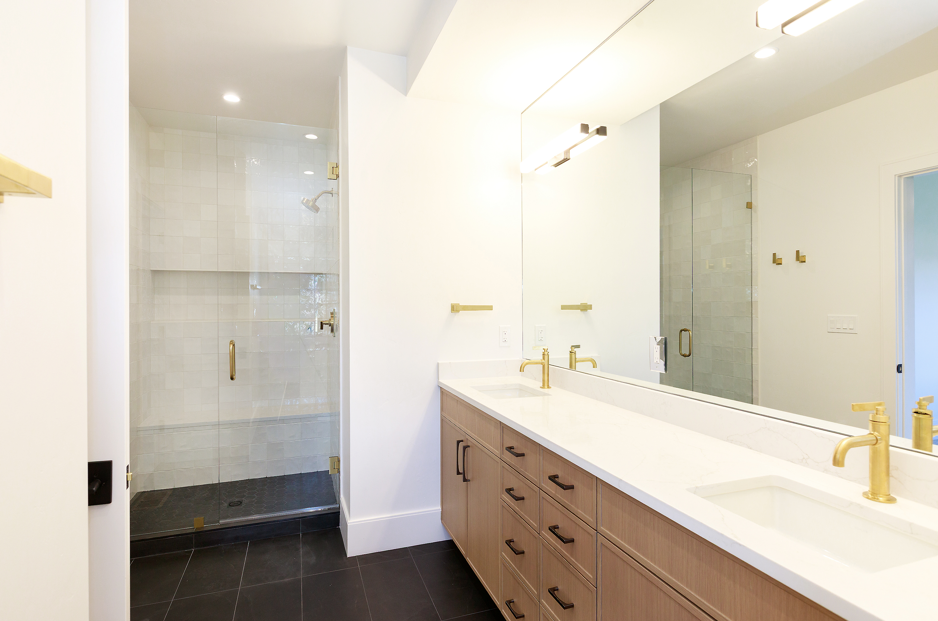 This is a bright and modern primary bathroom featuring a double vanity with a white countertop and light wood cabinetry. Gold fixtures add a touch of elegance, and a large mirror reflects the space, including a glass-enclosed shower with white tiled walls. The dark tile flooring provides a nice contrast to the light walls and fixtures.